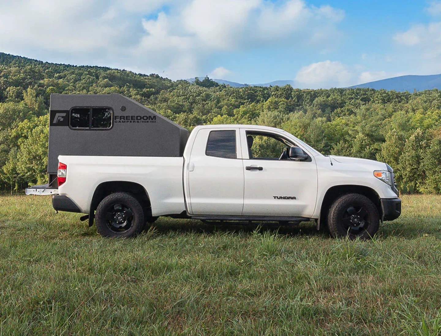 A white Toyota Tundra pickup truck with a black camper shell parked on a grassy field, surrounded by green trees with hills and a partly cloudy sky in the background.