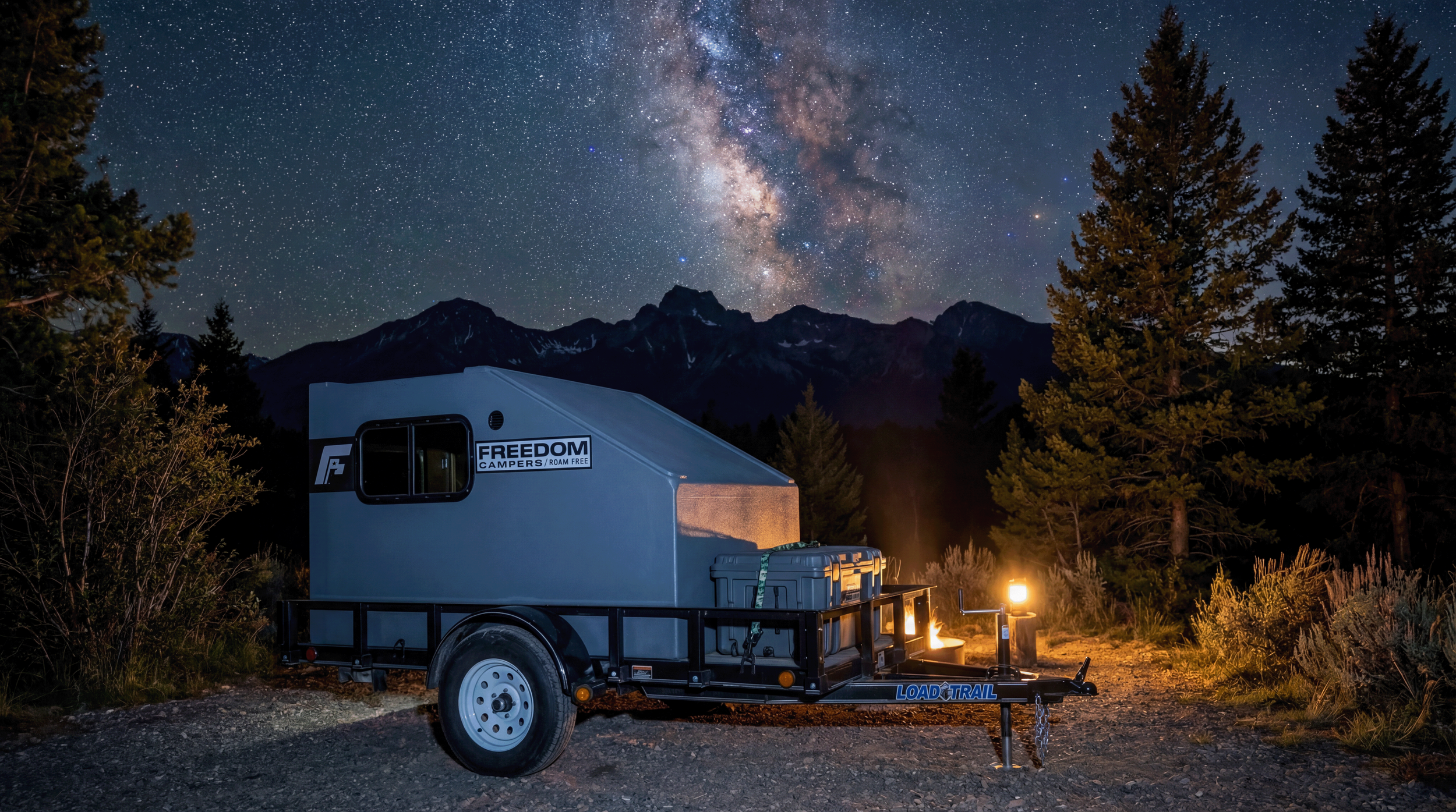 A compact travel trailer on a gravel campsite under a starry night sky with the Milky Way galaxy visible, surrounded by tall pine trees and mountains in the background, illuminated by a small campfire and a lantern.