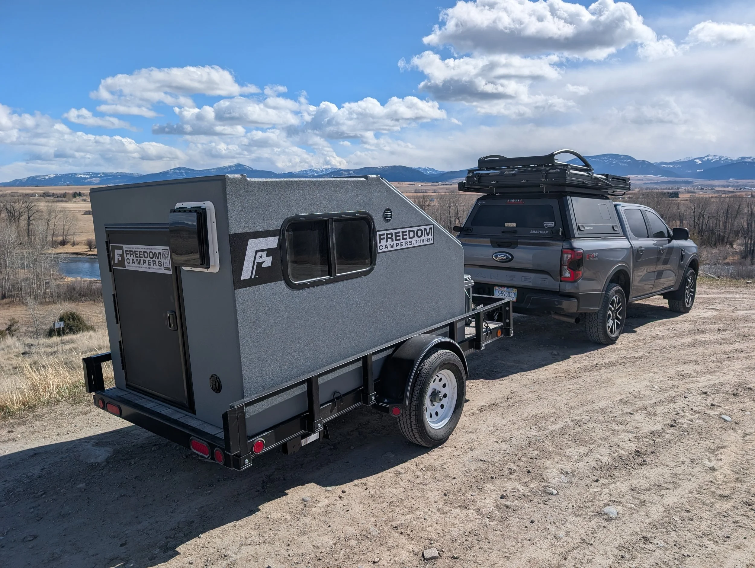 A gray Ford pickup truck attached to a small travel trailer with a gray exterior, parked on a dirt area overlooking a landscape with a river and snow-capped mountains in the distance under a partly cloudy sky.