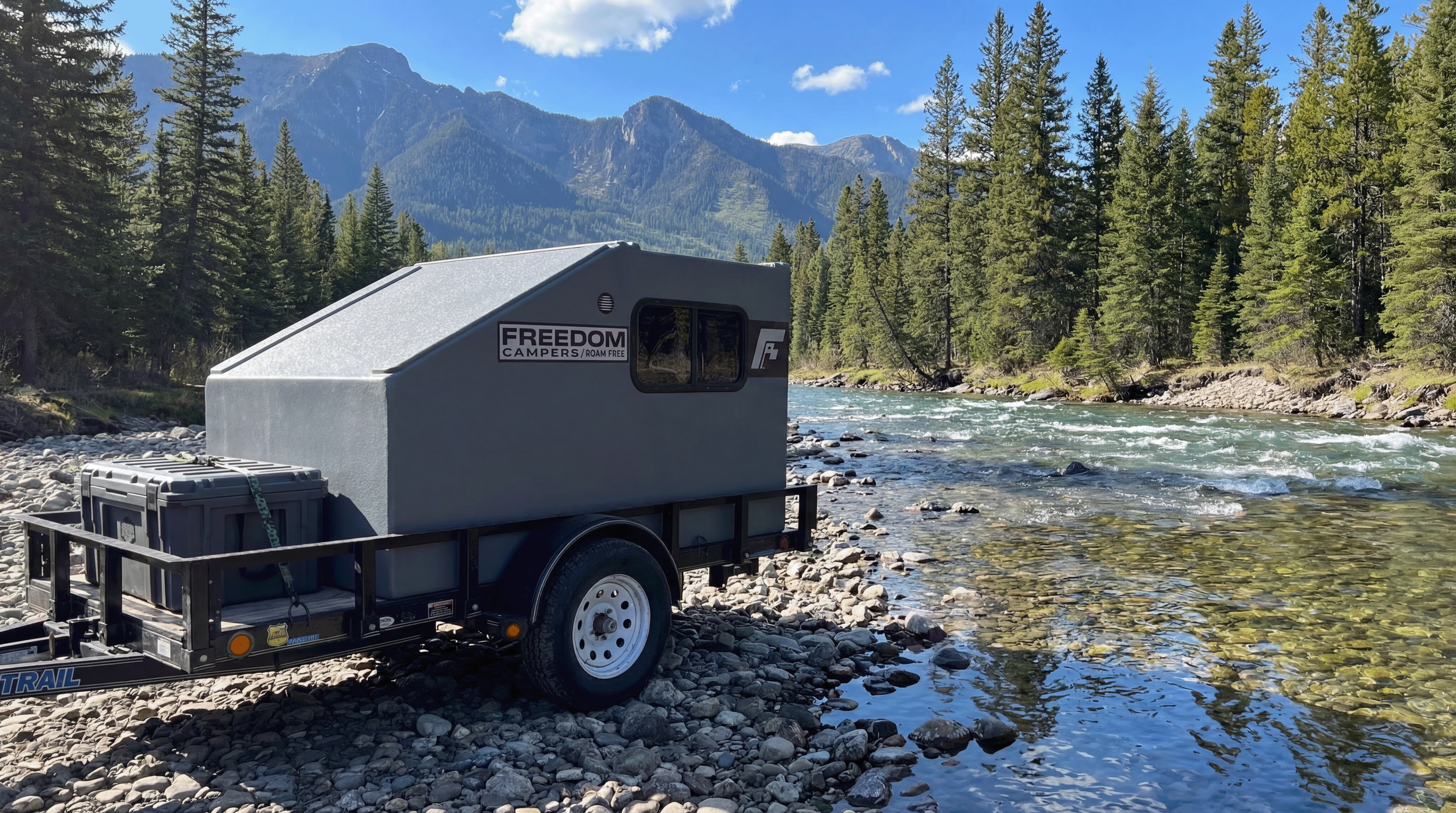 A gray camper trailer parked on a rocky riverbank in a forested mountainous area under a clear blue sky.