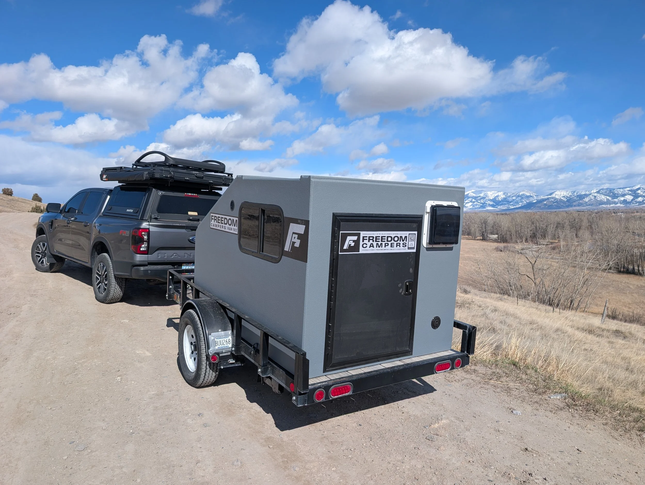 A gray camper trailer hitched to a black pickup truck parked on a dirt road in a rural area with mountains in the background.