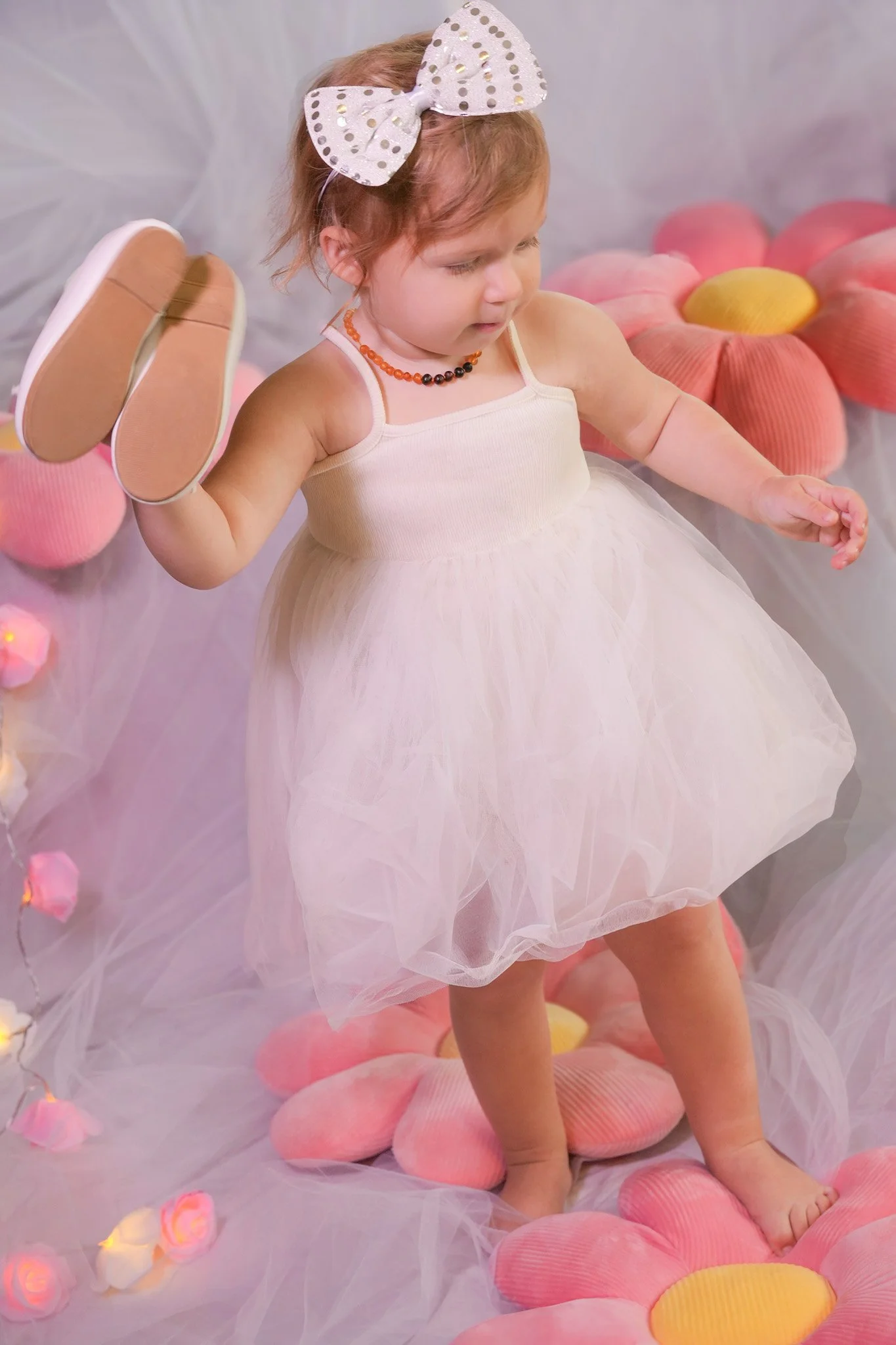 Baby girl wearing white dress, white hair band, holding her white shoes in one hand and starting to walk, On the background white fabric with pink flourish pillows and lights decorations.
