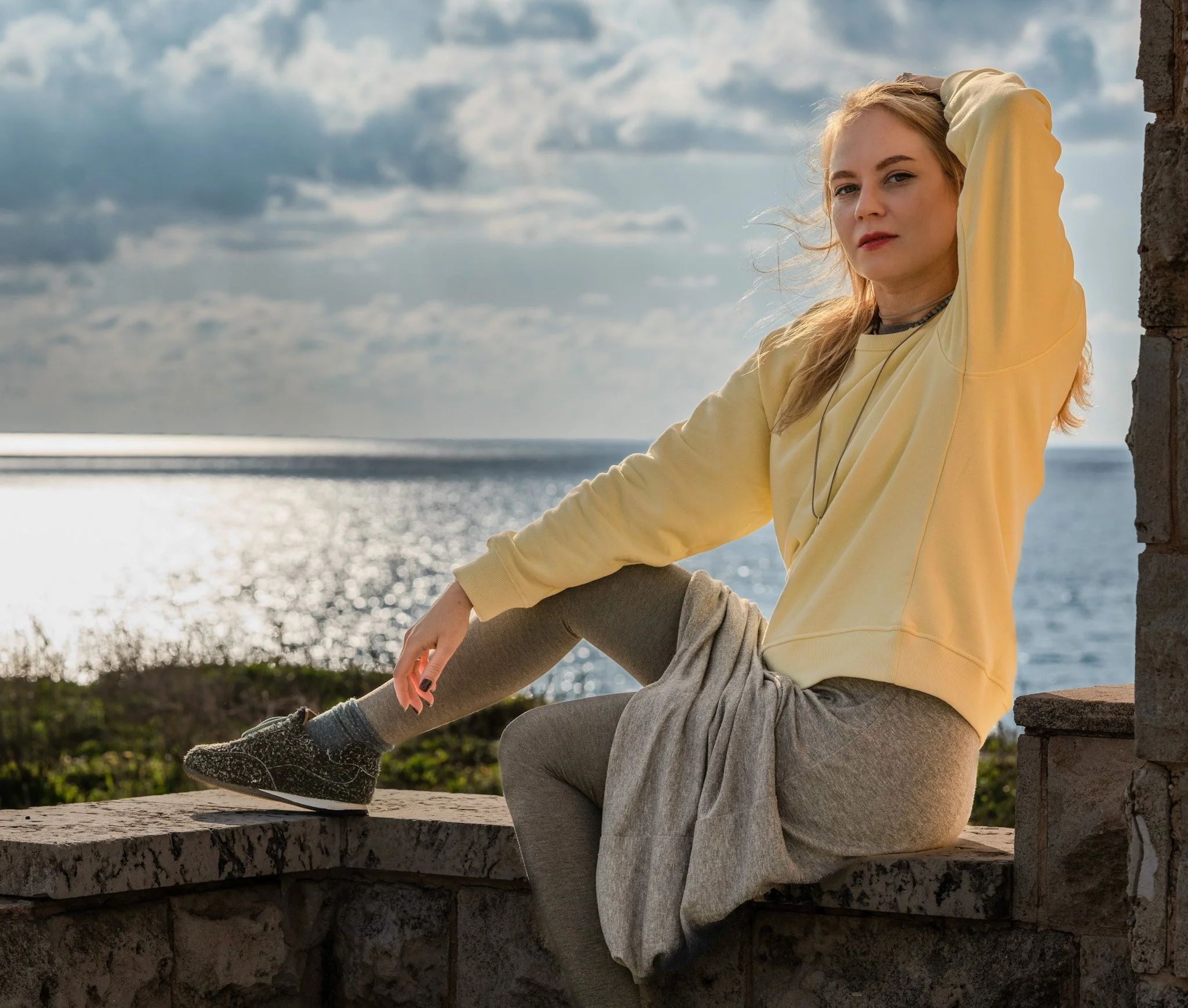 A young woman with blonde hair, wearing a yellow sweatshirt, gray leggings, and black sneakers, sitting on a stone wall by the water during sunset with a cloudy sky.
