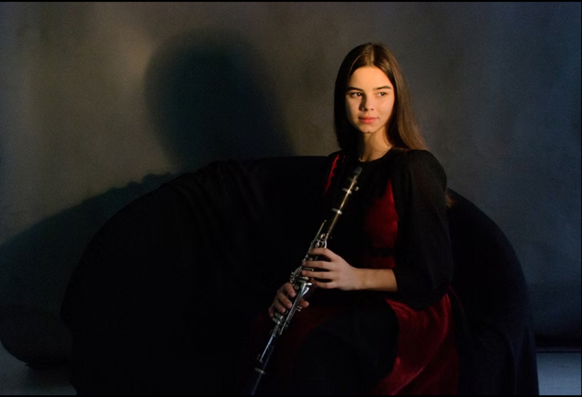 A young woman with long brown hair sitting on a black sofa holding a clarinet, dressed in a red dress and black shawl, with a dark, plain background.