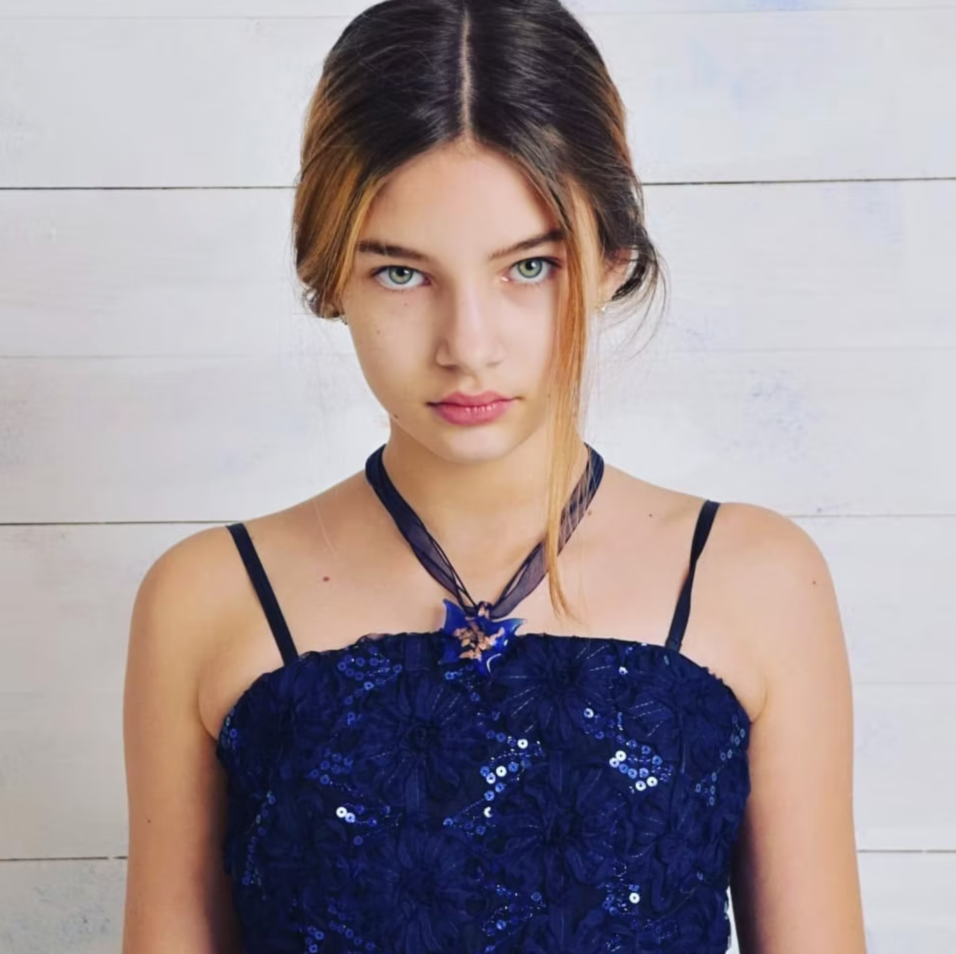 A young woman with light skin, green eyes, and long hair styled with a side part, wearing a navy blue dress with sequins and thin spaghetti straps. She is standing against a white wooden wall.