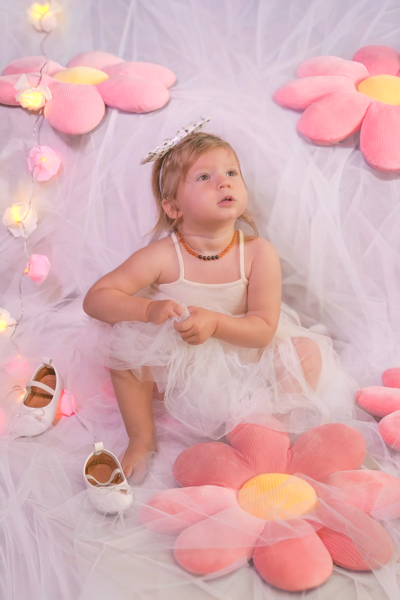 Baby girl wearing white dress, white hair band, and white shoes near her, On the background white fabric with pink flourish pillows and lights decorations.