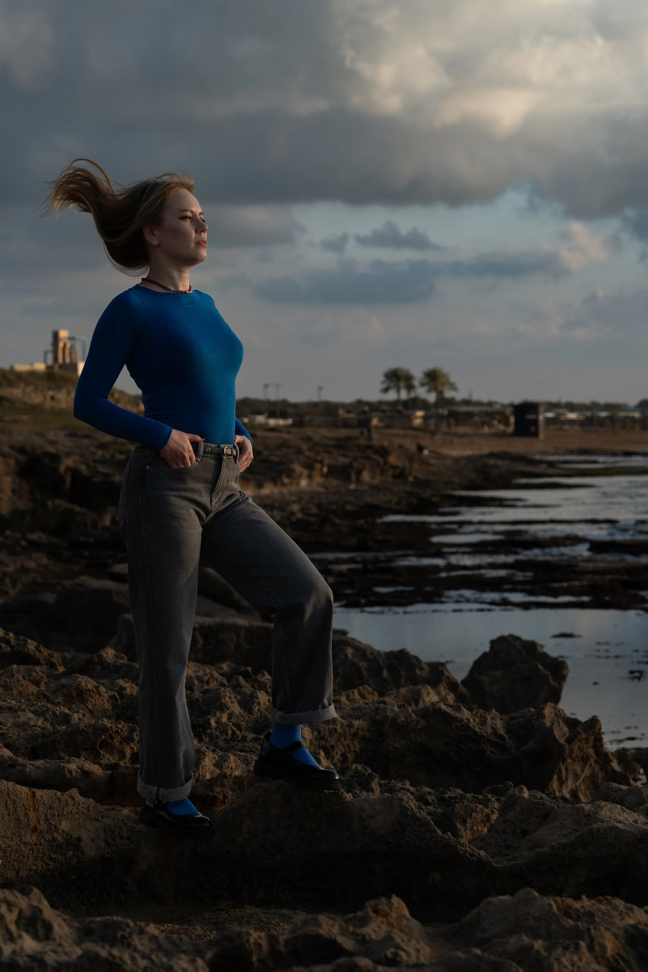 A woman with long hair wearing a blue long sleeve shirt, gray jeans, and black shoes stands on a rocky shoreline with her hands in her pockets, under a cloudy sky, near the ocean with small waves, in a coastal  setting at sunset .