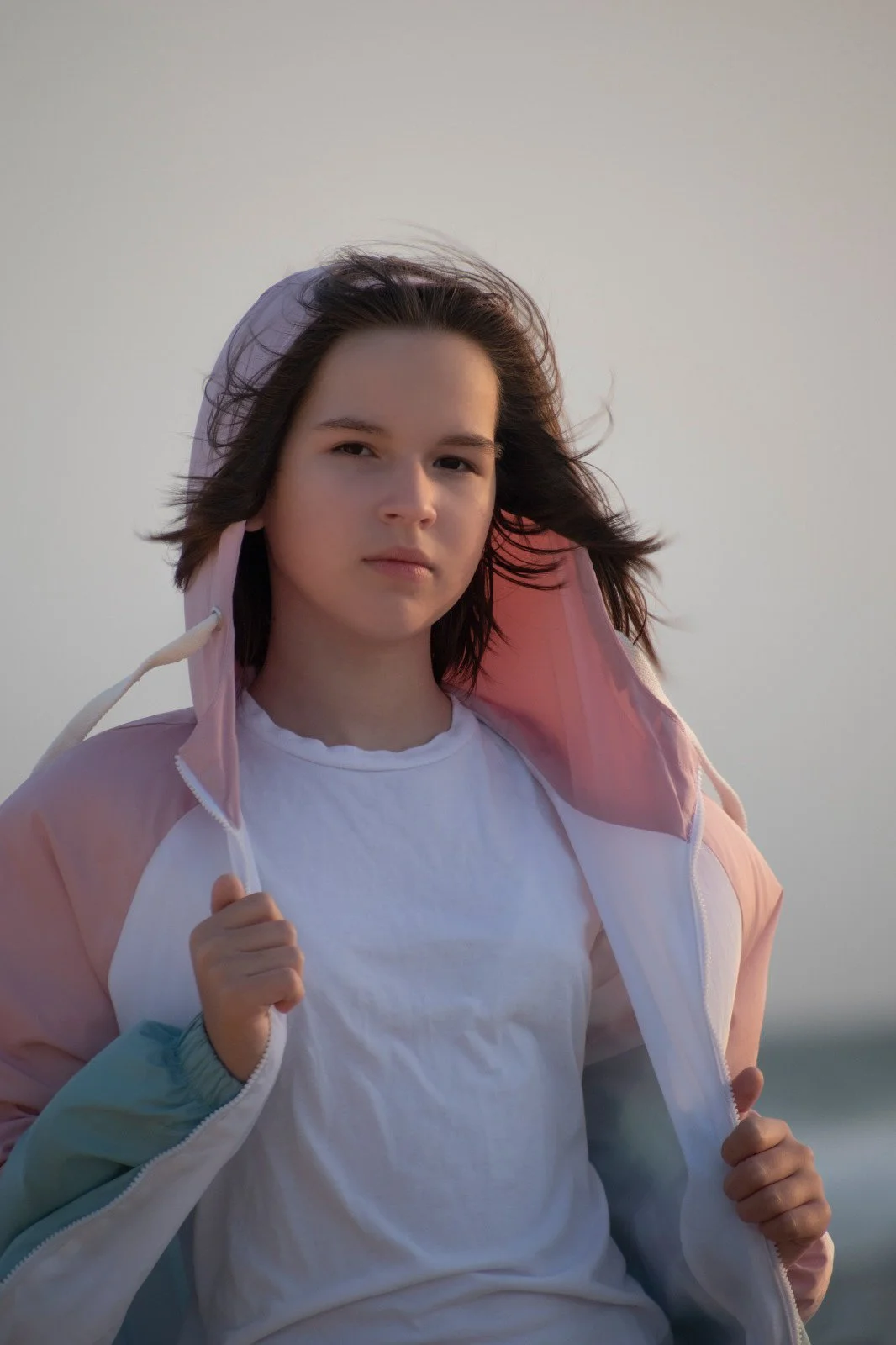 A young woman with shoulder-length brown hair wearing a white T-shirt and a pink and light blue windbreaker holding the open front of her jacket, standing outdoors with a neutral background.