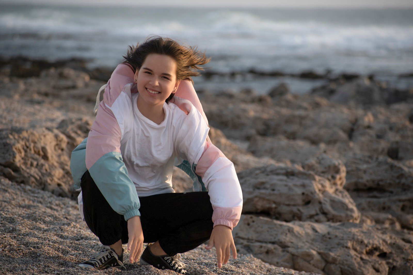 A girl crouching on a rocky beach, smiling, with the ocean in the background.