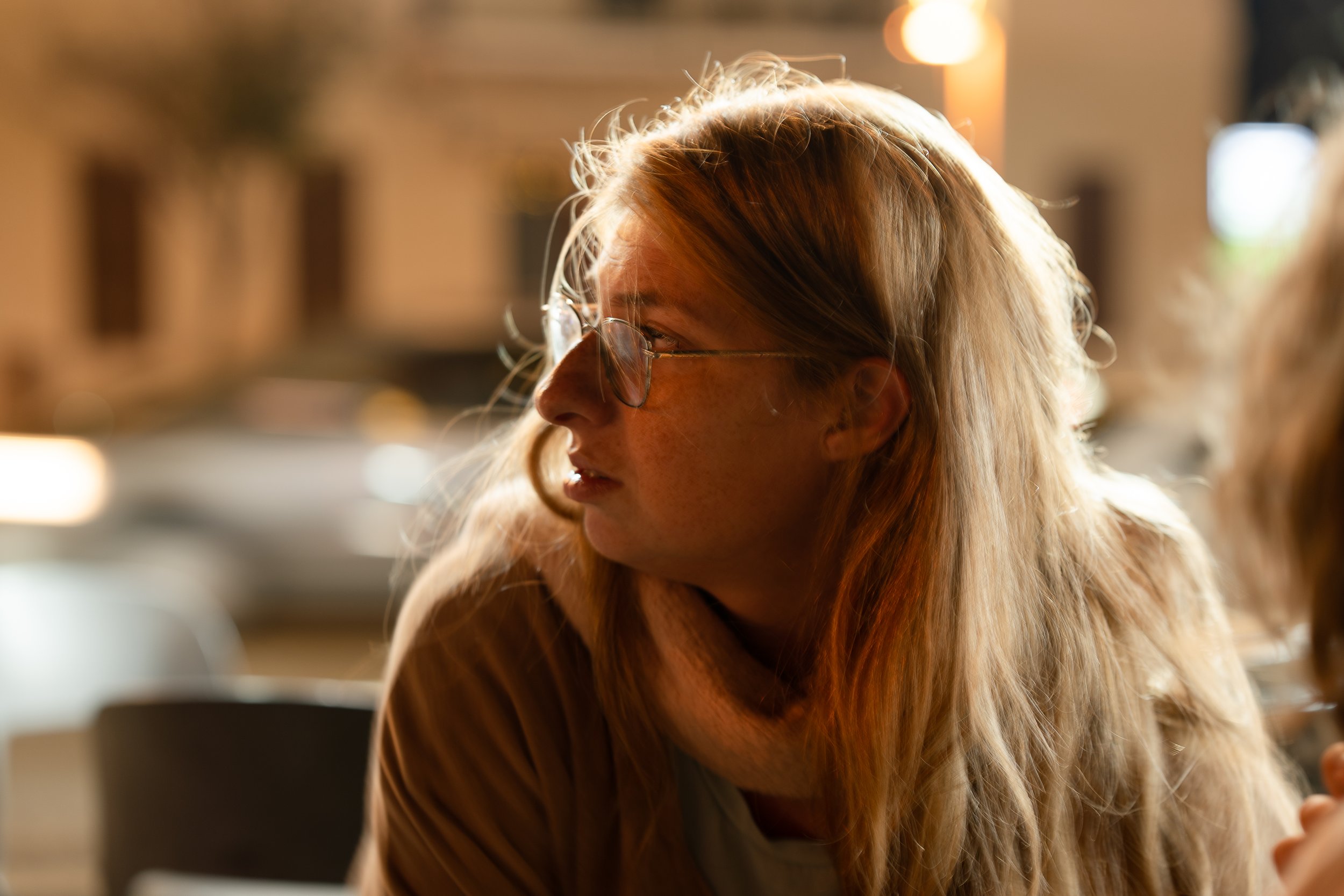 A woman with long red hair wearing glasses, looking to her left, illuminated by soft warm lighting in an indoor setting.