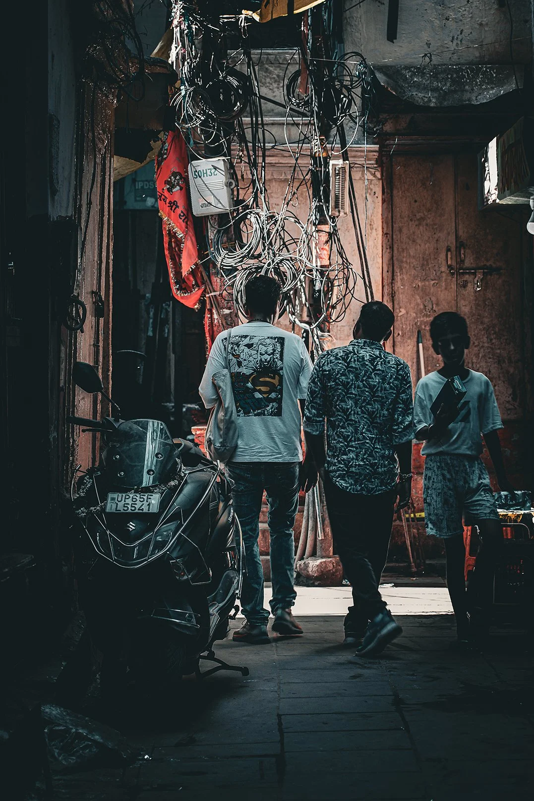 Three young men walking through a narrow alleyway in Kashi with tangled electrical wires overhead. One is carrying a bag, and they are near motorcycles and a wall with a door, in an urban setting.