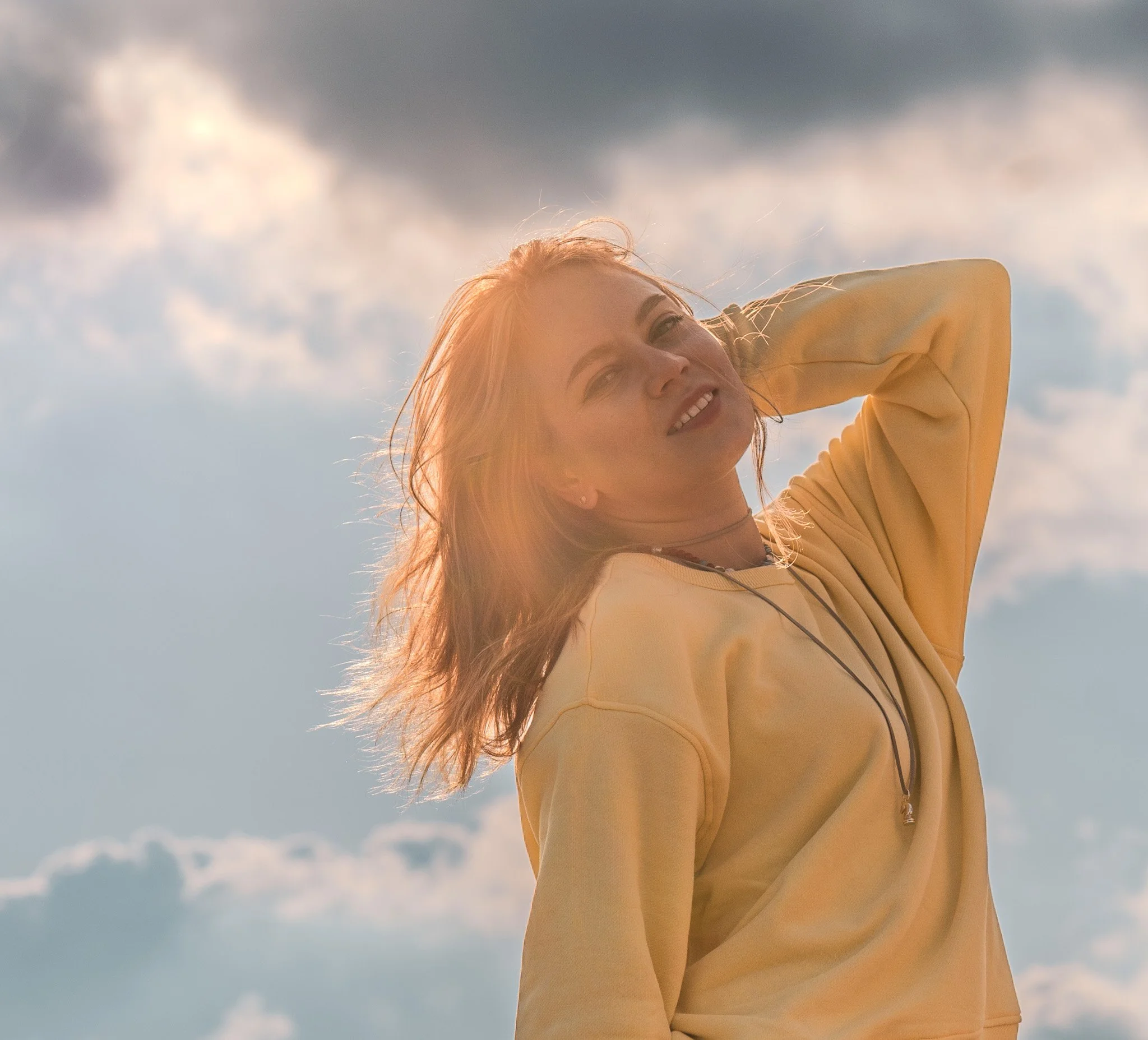 A woman with blonde hair smiling and looking at the camera, wearing a yellow sweatshirt, outdoors with a cloudy sky in the background.