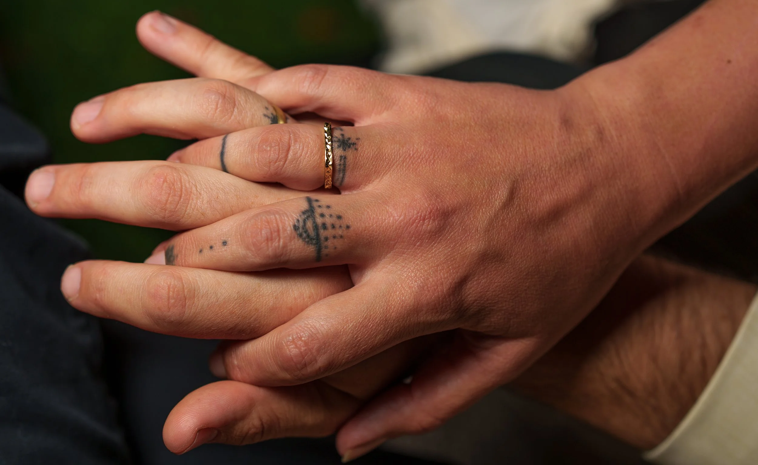 Close-up of two hands gently clasped together, one with tattoos and wearing a wedding rings.