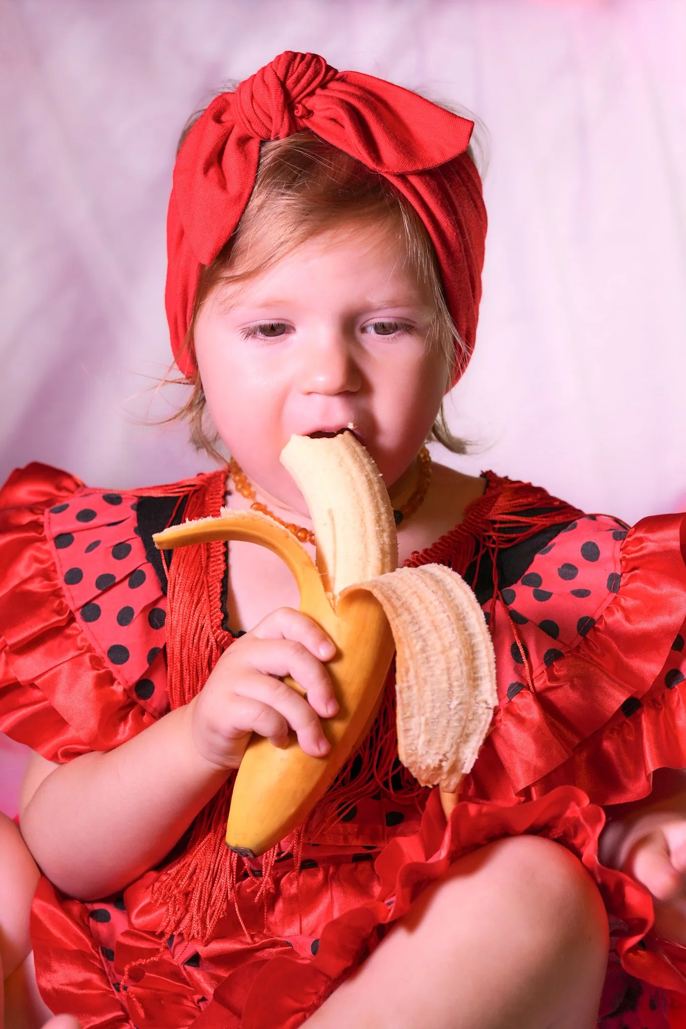 Cute baby girl in a red dress and red hair band eating banana