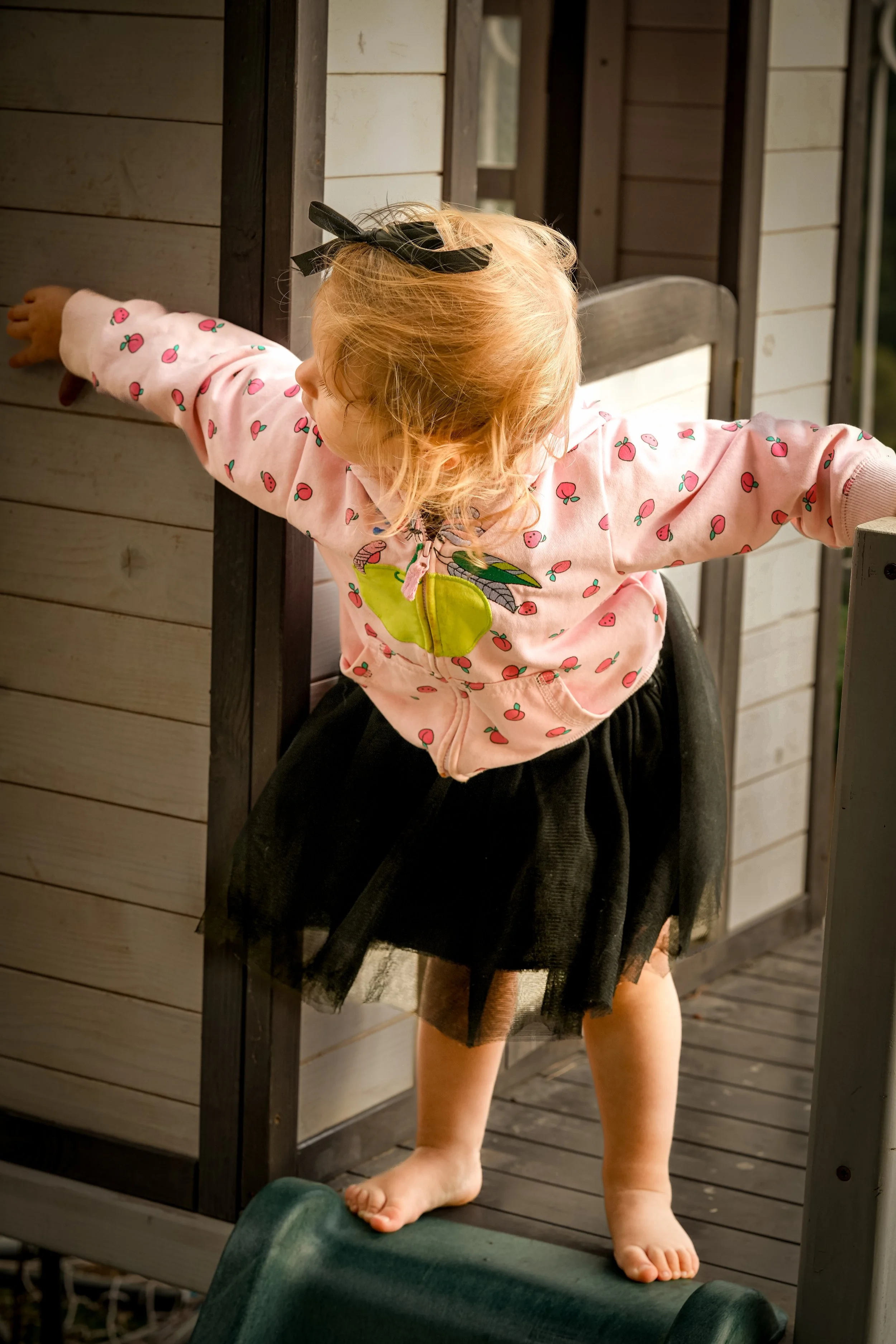 A young girl with curly red hair, wearing a pink hoodie with a fruit print and a black skirt, balancing on a green stares outdoors near a toy house, with her arms outstretched.