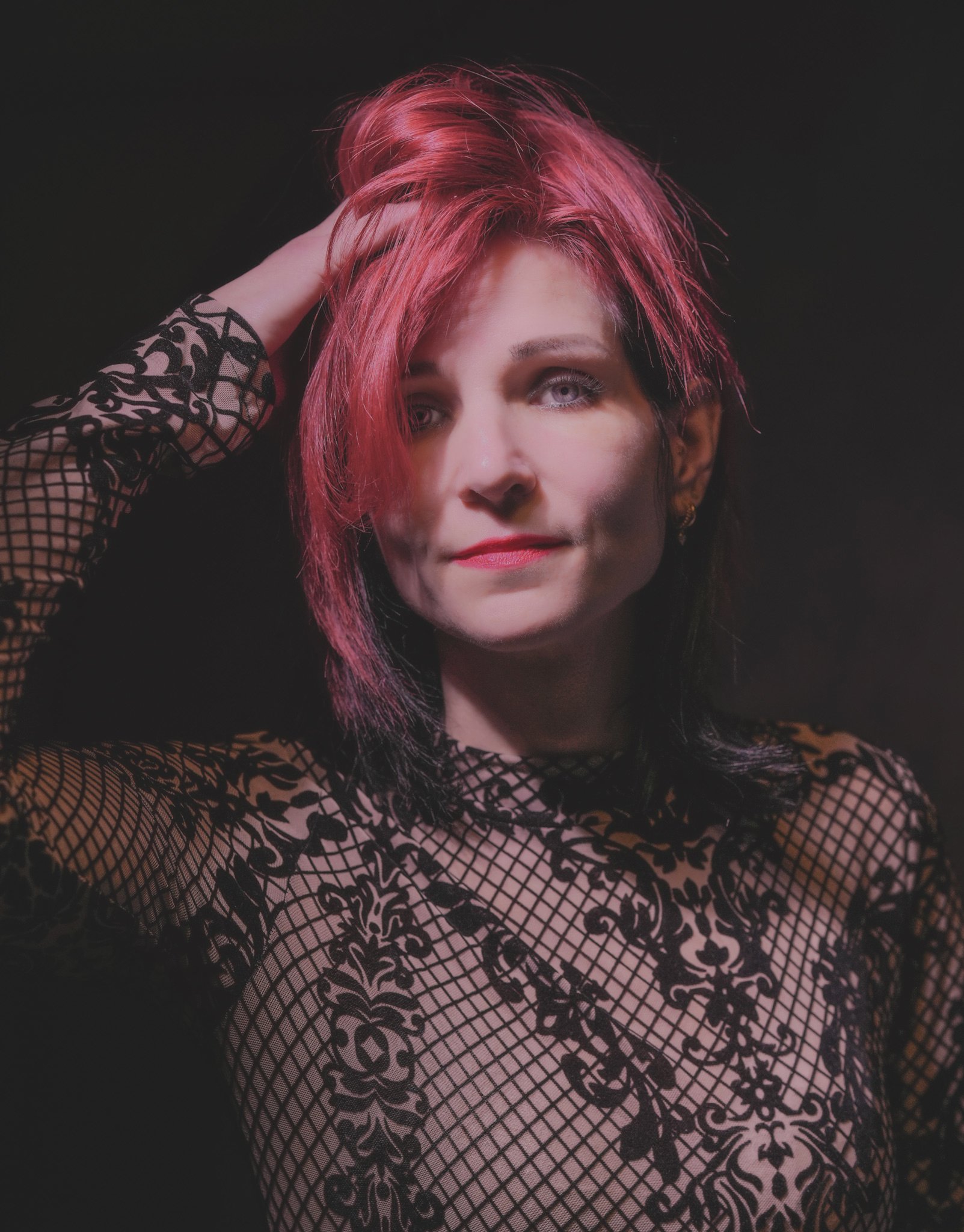 Portrait of a woman with pink hair and blue eyes, wearing a black lace top, posing with her hand on her head against a dark background.