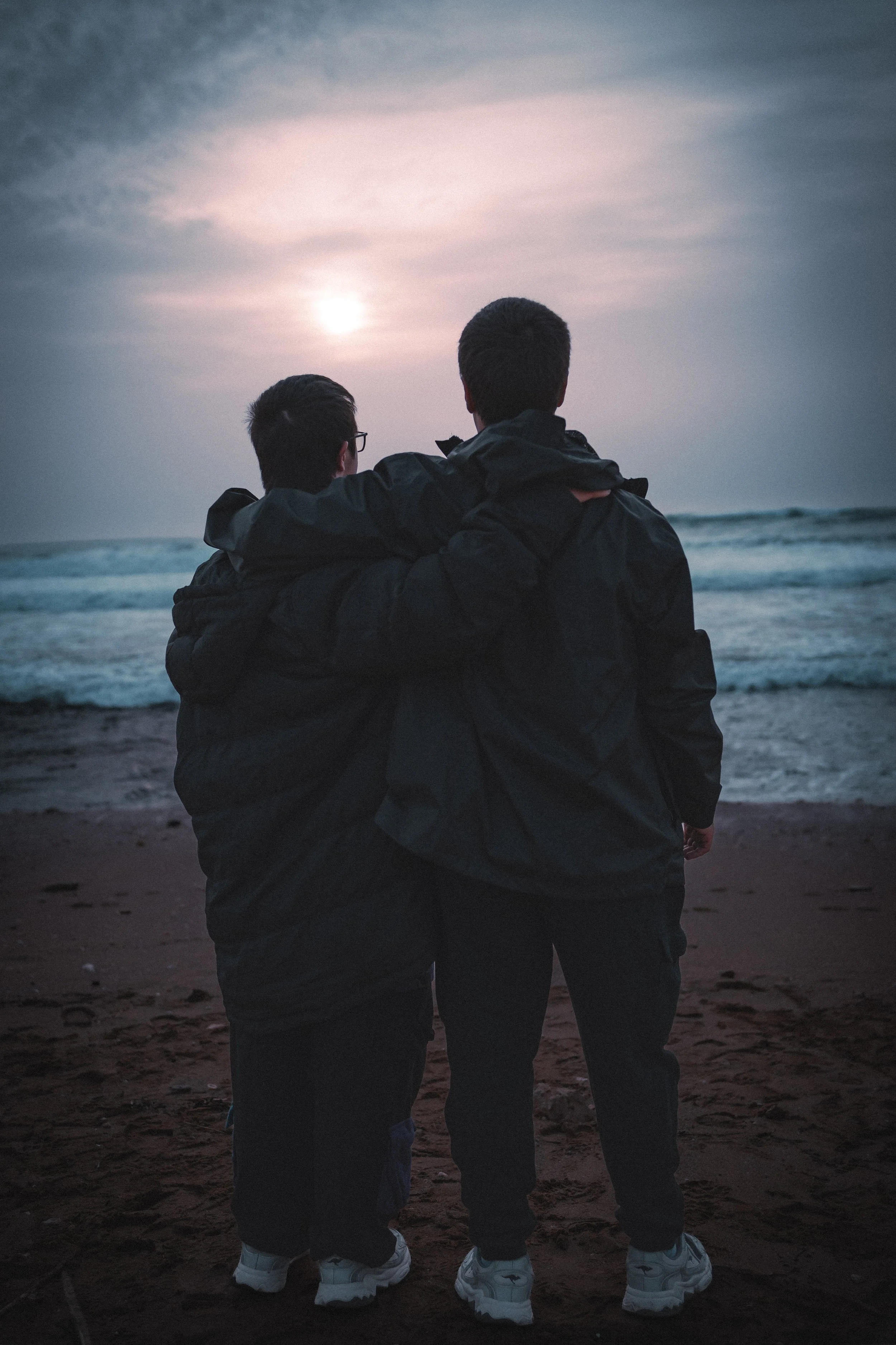 Two brothers standing on a beach, with one arm around each other's shoulders, watching the sunset over the ocean.