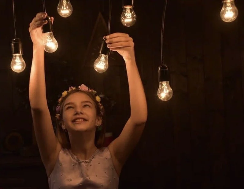 A young girl with a floral crown smiling and reaching up to touch hanging light bulbs in a dark room.