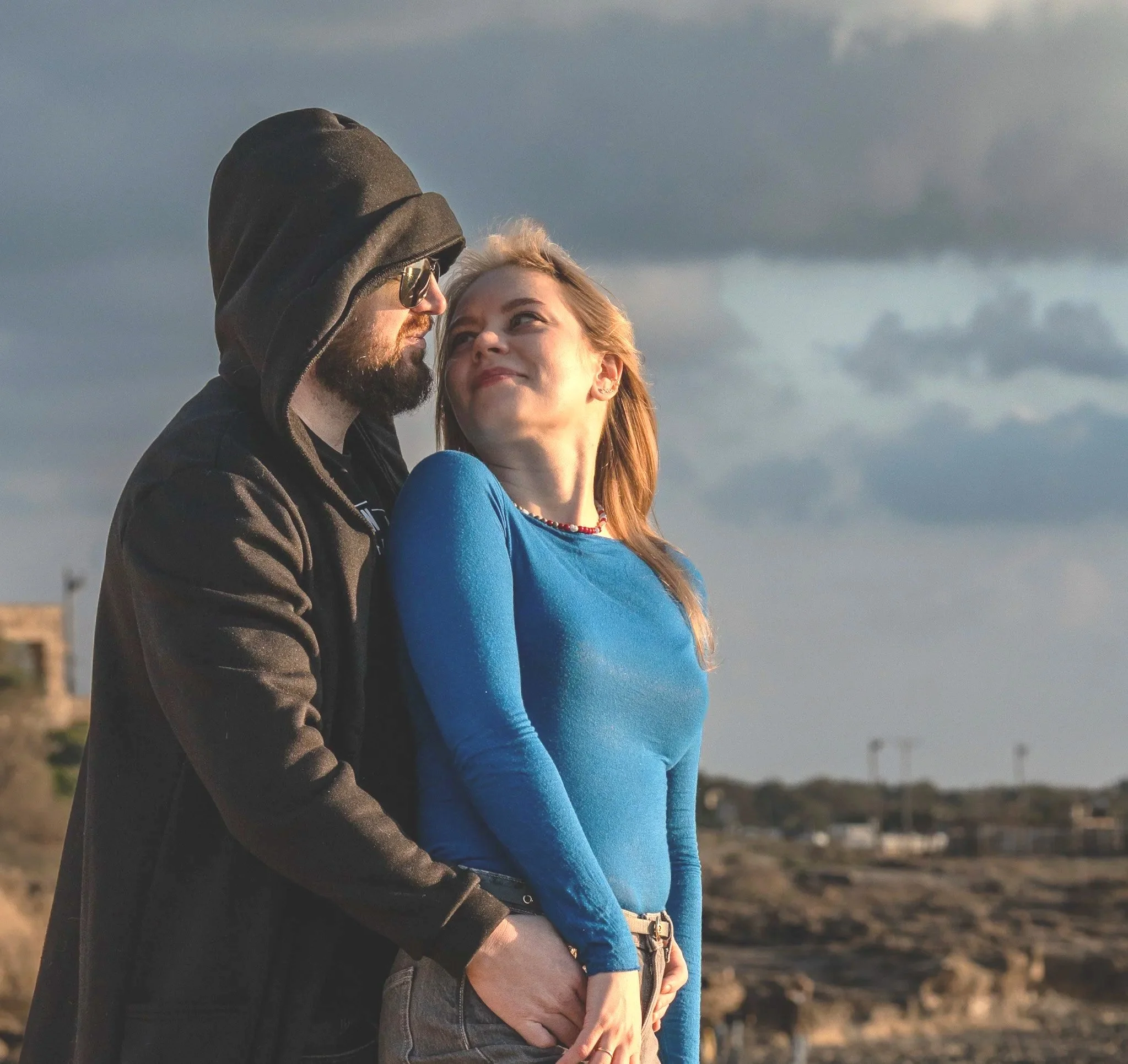 A man with a beard, sunglasses, and a hoodie embraces a woman in a blue long-sleeve shirt outdoors, with a cloudy sky and distant landscape in the background.