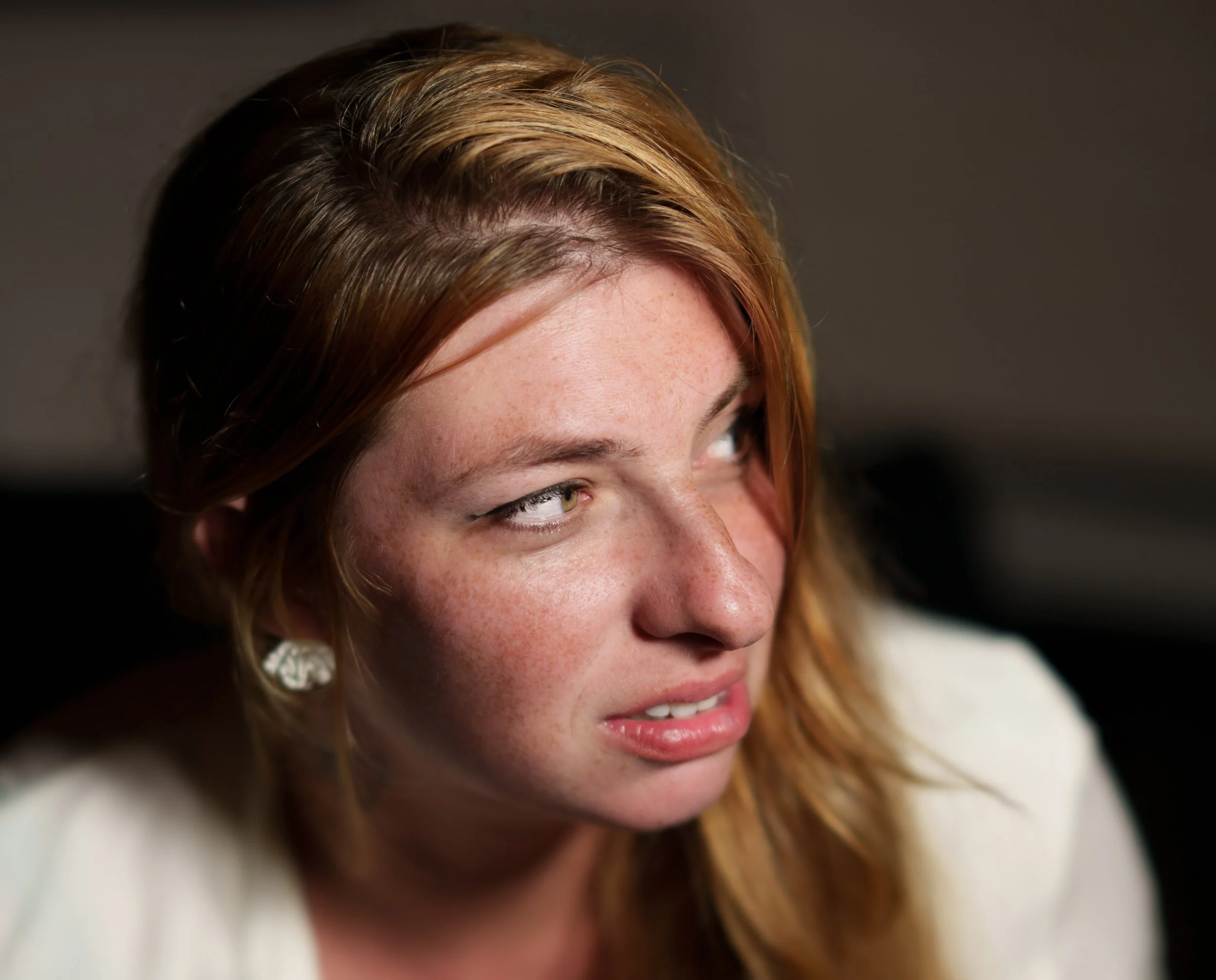 Close-up of a woman with reddish-brown hair, freckles, and light makeup, looking to her right with a slight smile, against a dark background.