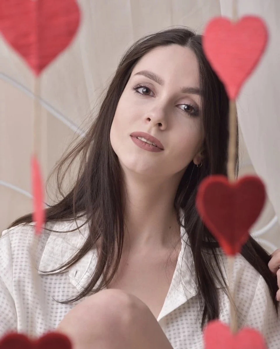 A young woman with long dark hair and light makeup looks into the camera through a string of red heart-shaped decorations.