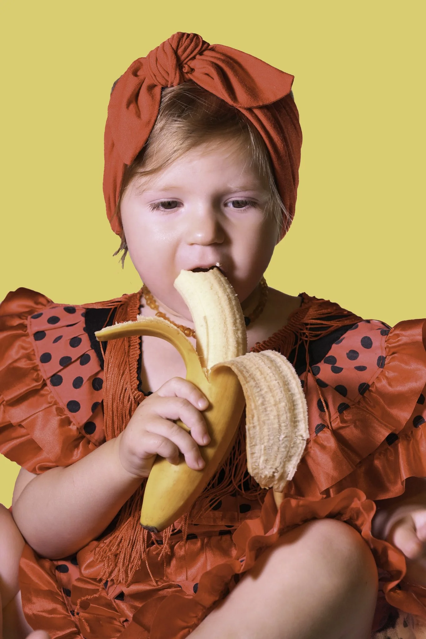 A young girl in a red polka dot dress and matching head wrap is eating a peeled banana against a yellow background.