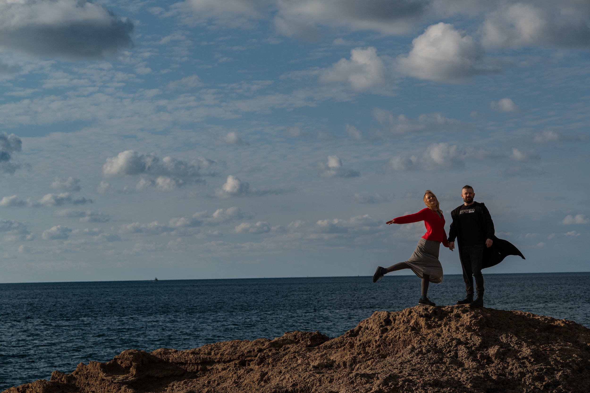 A couple standing on a rocky shoreline by the ocean with a sky filled with scattered clouds in the background.