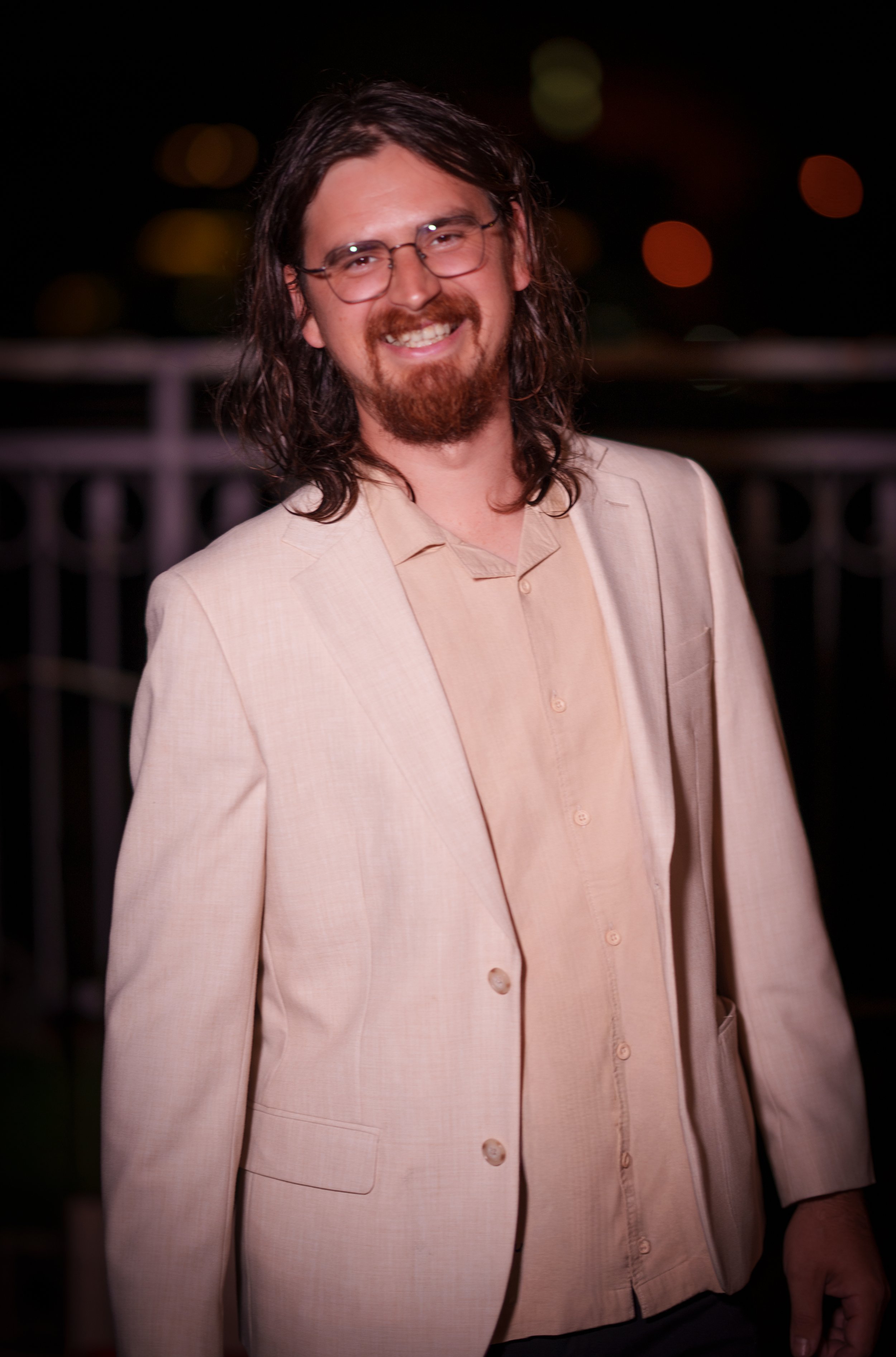 Man with long hair, glasses, and beard smiling, wearing a beige suit jacket and shirt, at night with out-of-focus lights in the background.