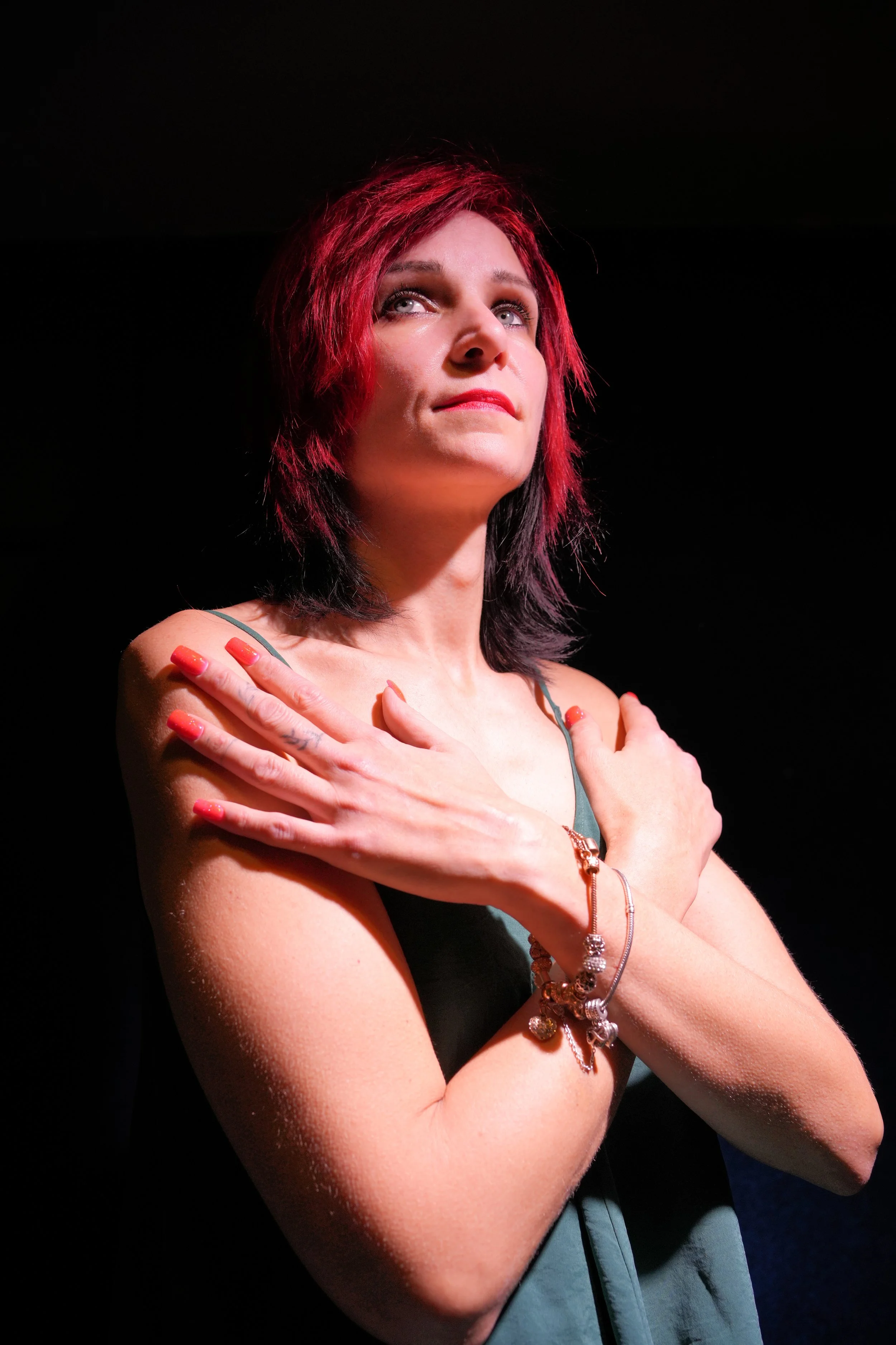 Stage scene. A woman with red and black hair standing against a black background, with her arms crossed over her chest, wearing a sleeveless dress and jewelry, gazing upward.