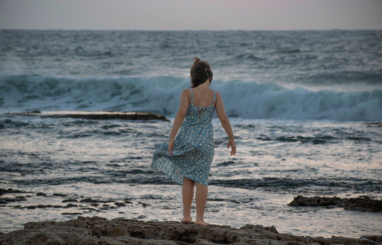 A girl in a blue floral dress walking barefoot on rocks at the beach with the ocean and waves in the background.