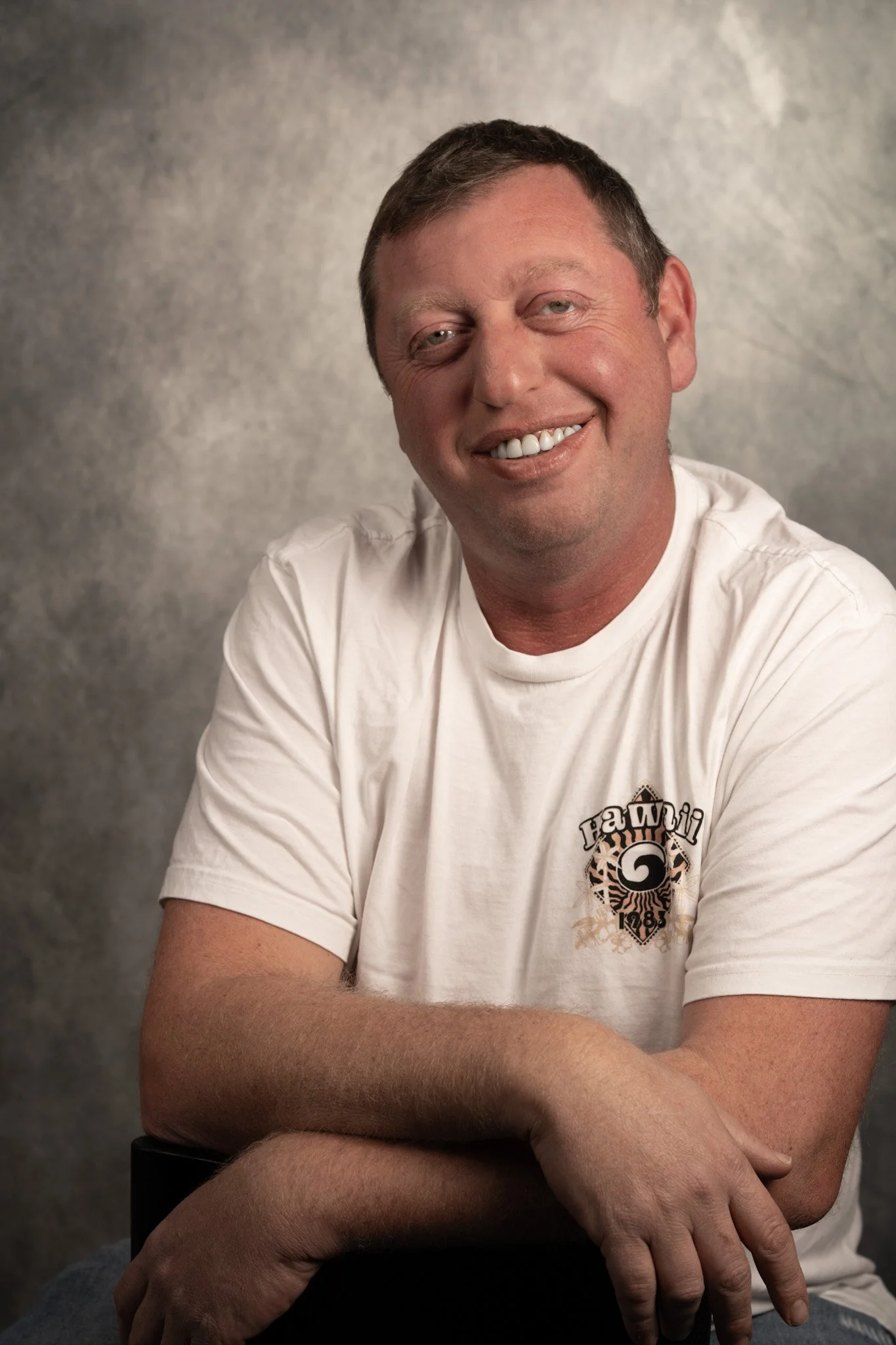 A smiling man with short brown hair and light skin, wearing a white T-shirt with a logo on it, sitting with his arms crossed against a gray studio background.