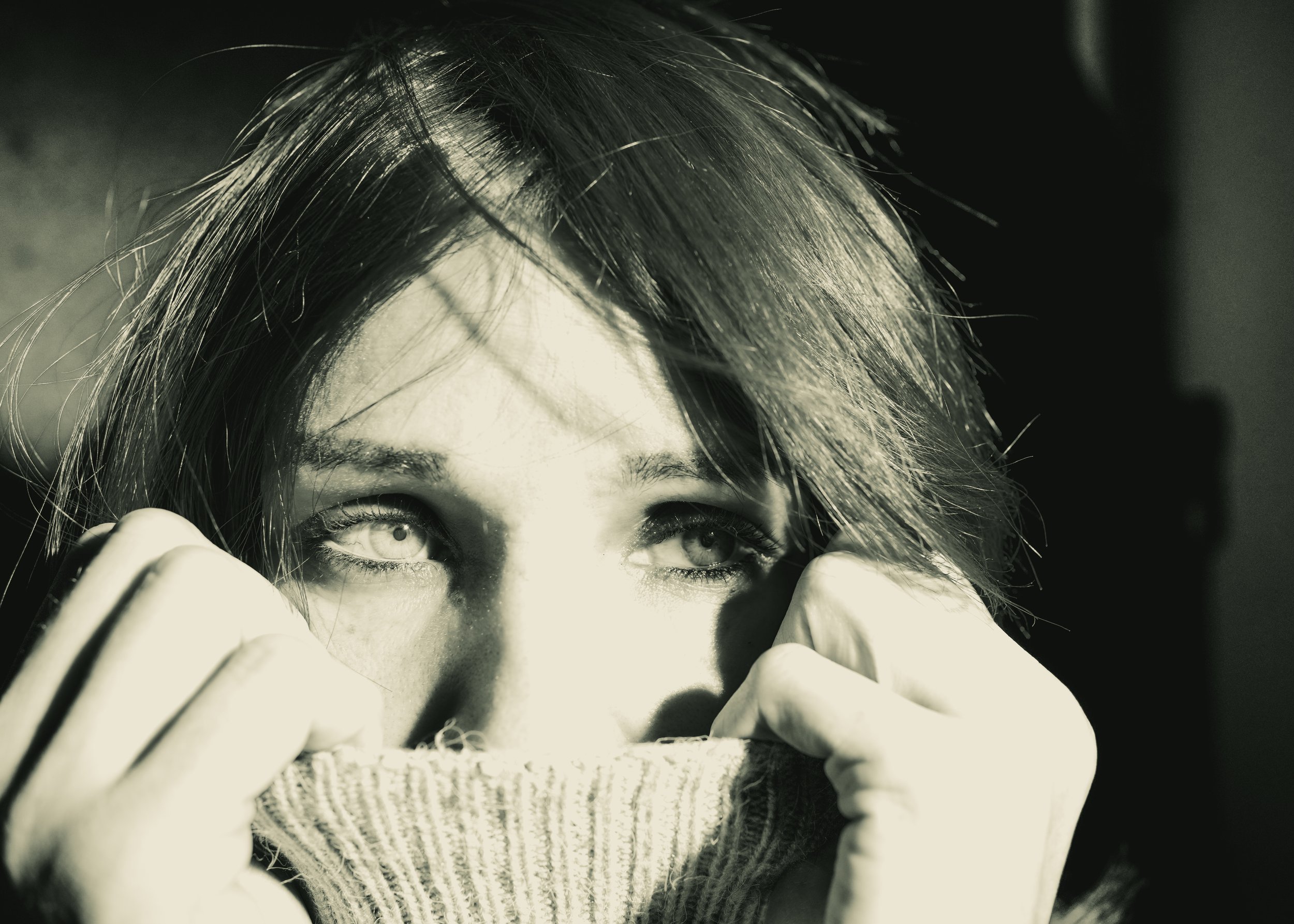 Close-up of a woman with light-colored eyes holding a sweater over her mouth, with sunlight casting shadows on her face.
