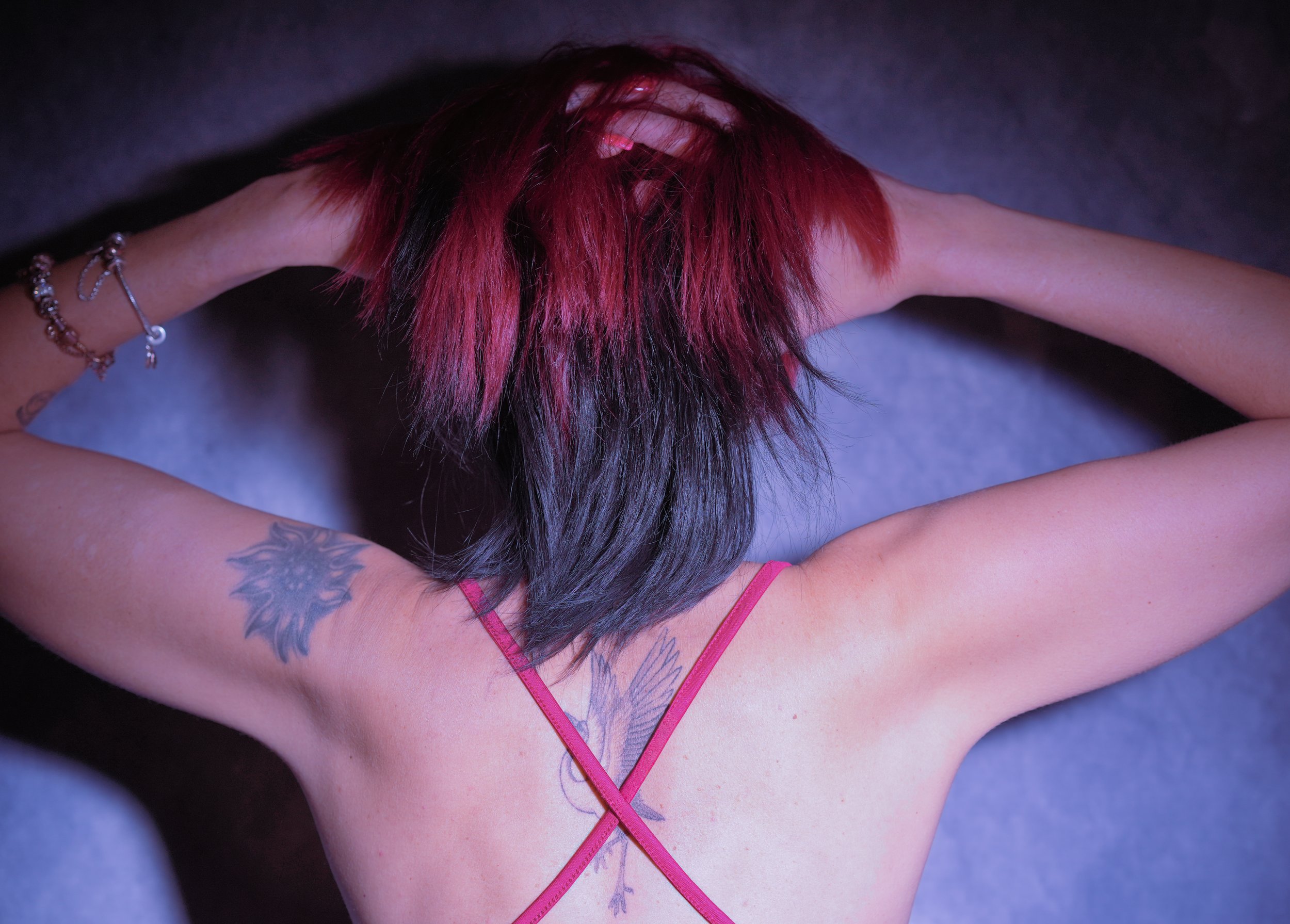 A woman standing, showing  her back with her arms behind her head, showing her tattooed back and red and black hair, wearing a pink strappy top and bracelets.