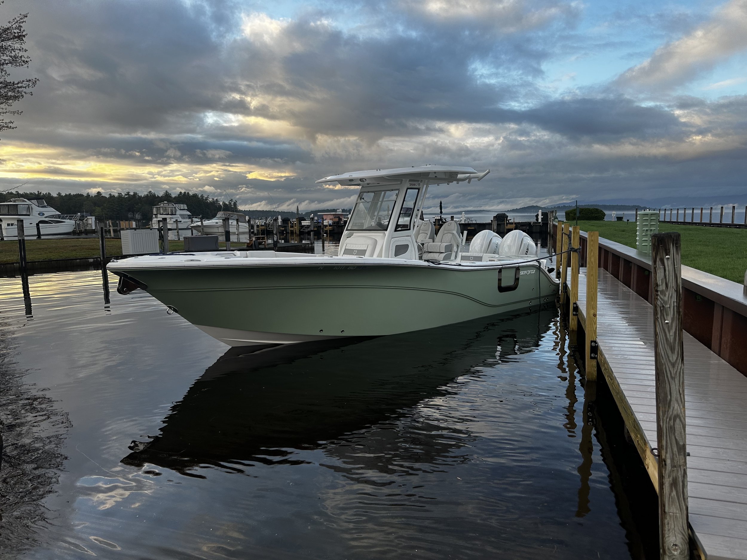 A boat docked at a marina with a cloudy sky overhead and calm water reflecting the scene. Multiple boats are visible in the background, docked along the pier.