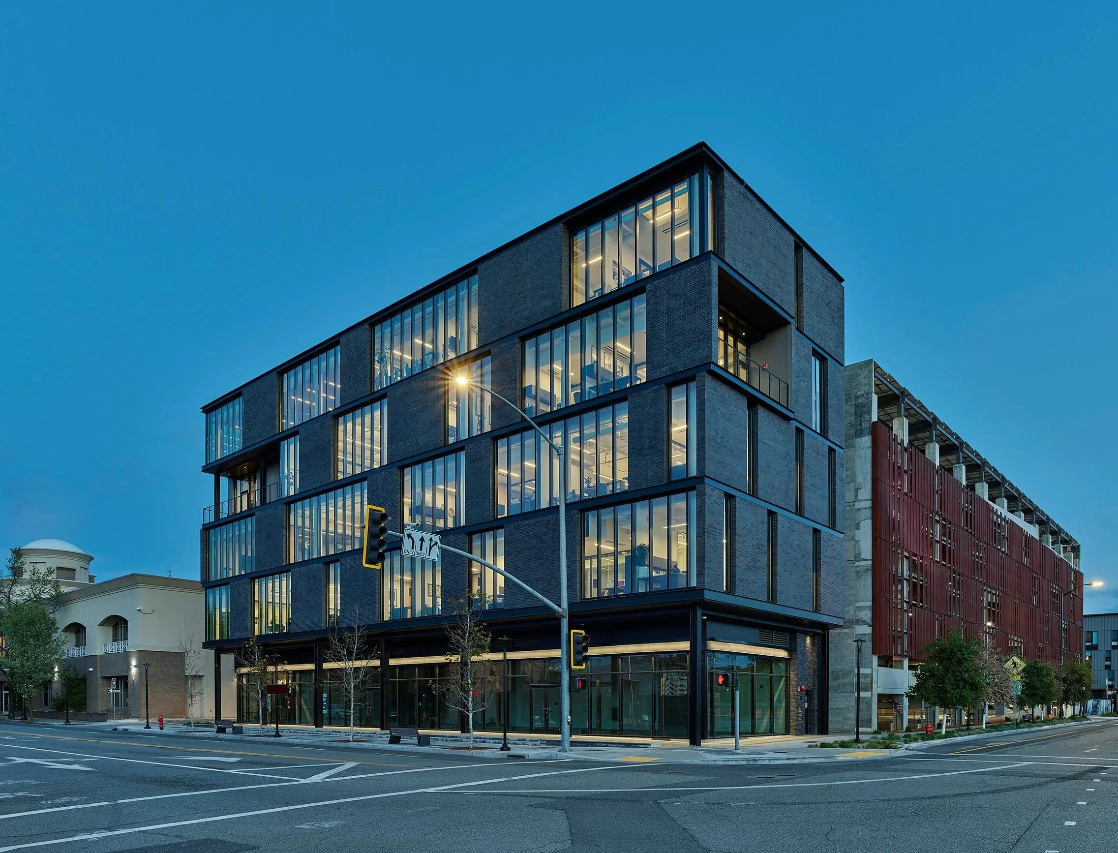 Shasta College in Redding, California. Part of Block 7 Development. A modern multi-story building with large glass windows and a brick exterior, located at an intersection with traffic lights, street signs, and nearby trees, during dusk.