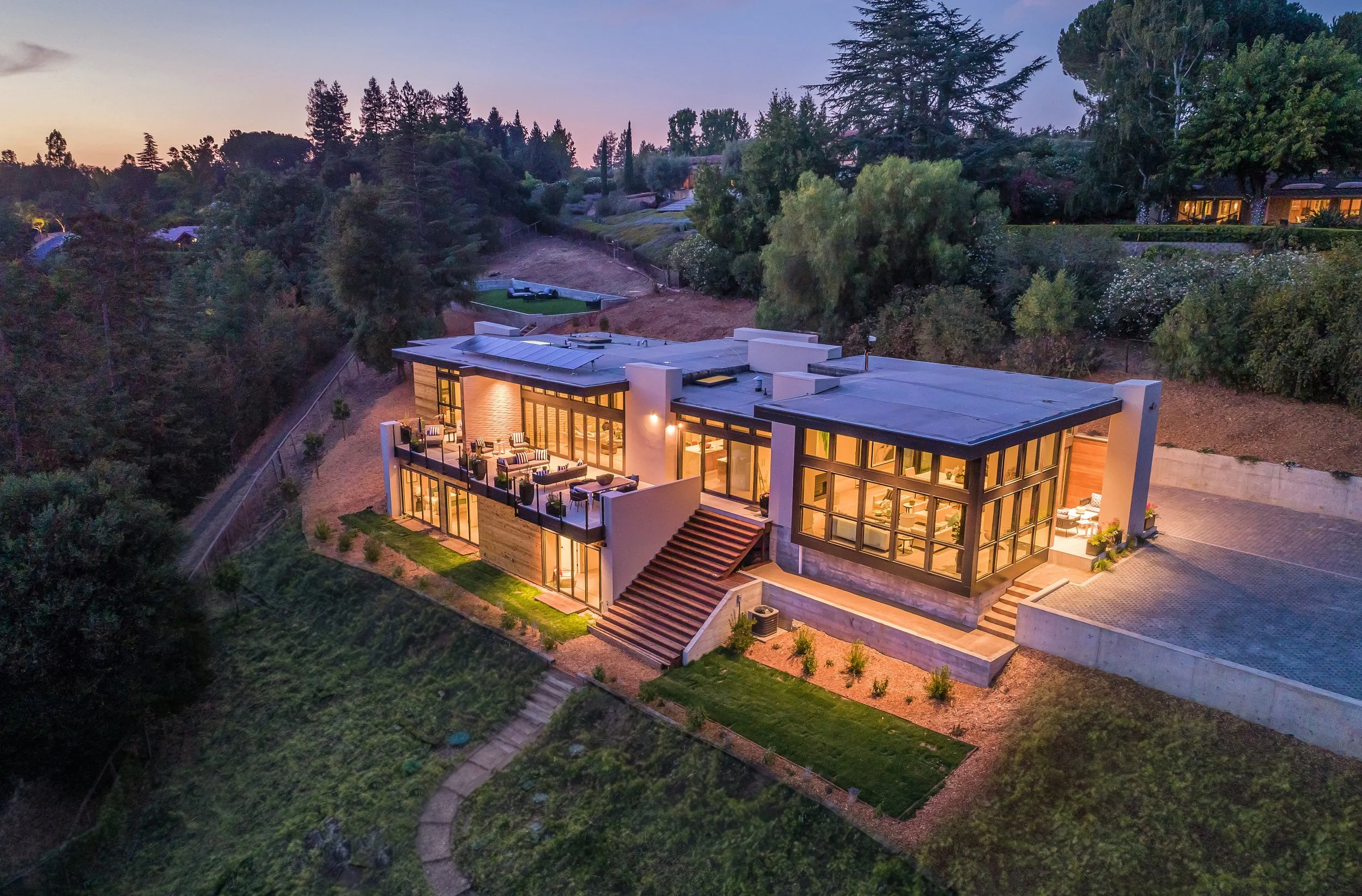 Los Altos Hillside Home in Los Altos, California. Modern house with large glass windows, illuminated interior, overlooking a hilly wooded landscape at dusk with trees and a garden.