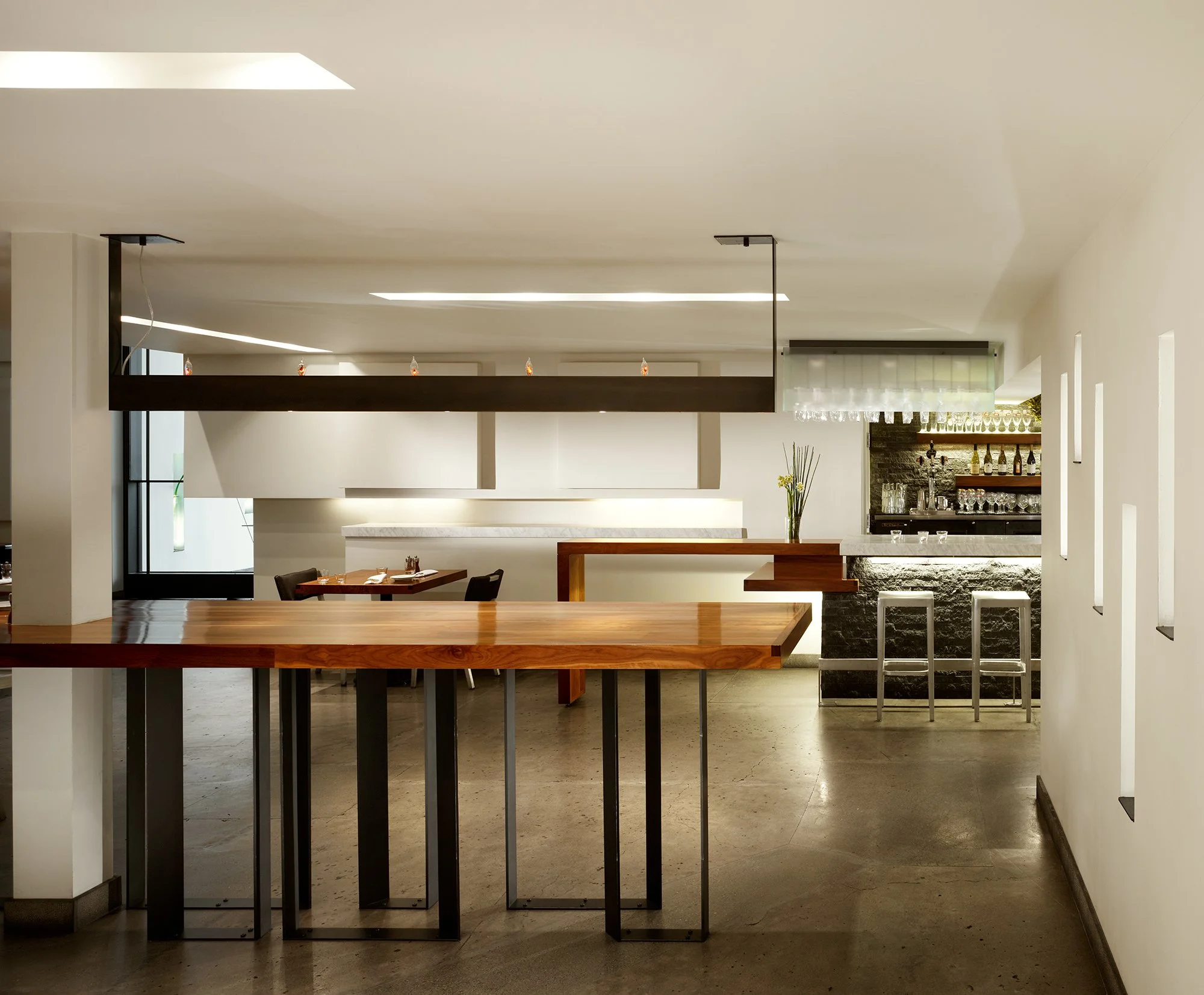 Local Wine Merchant restaurant and bar in San Francisco. Custom fabricated dining area with a long dark hanging light fixture, and a bar in the background with bottles and glasses, featuring minimalist decor and neutral colors.