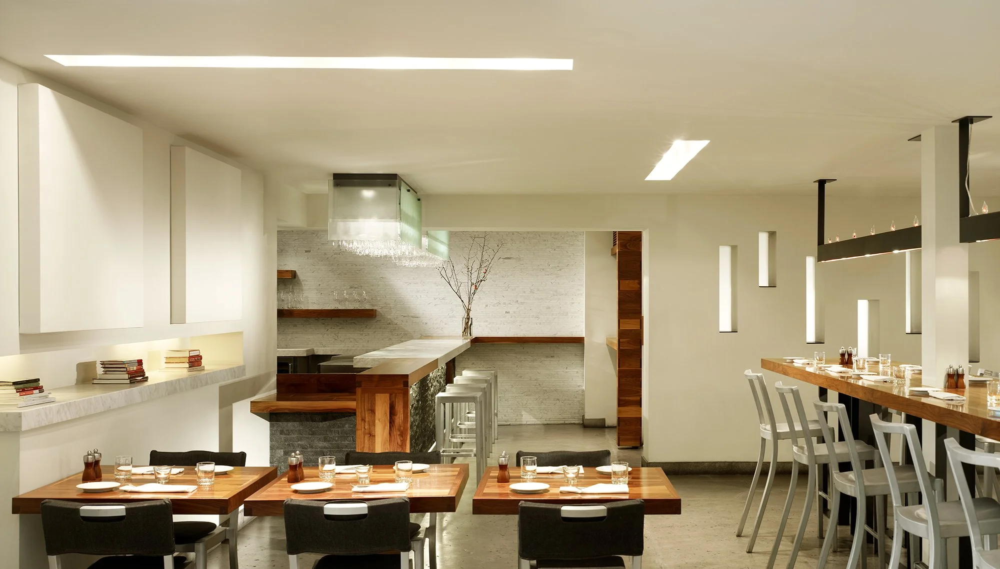 Local Wine Merchant restaurant and bar in San Francisco. Custom fabricated interior of a modern restaurant with wooden tables, chairs, and minimalistic decor, including a white brick wall and small, narrow windows.