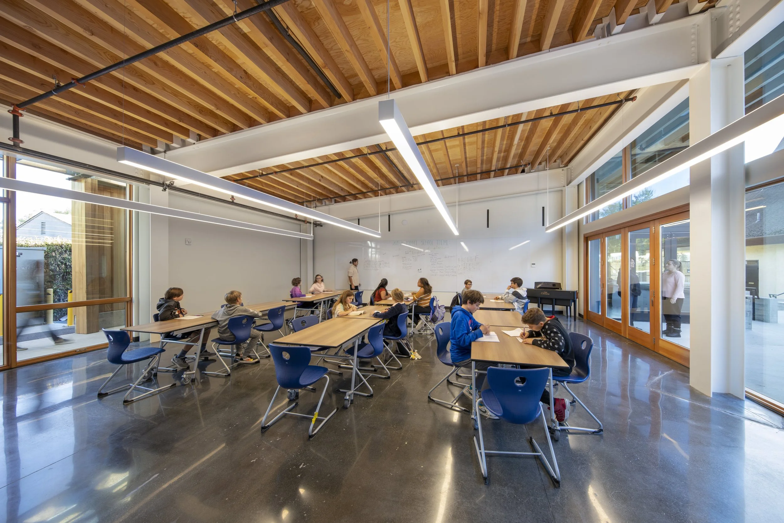 Spring Hill Montessori School in Petaluma, California. A classroom with students seated at tables, a teacher standing in front of a whiteboard, large windows letting in natural light, wooden ceiling beams, and modern lighting fixtures.