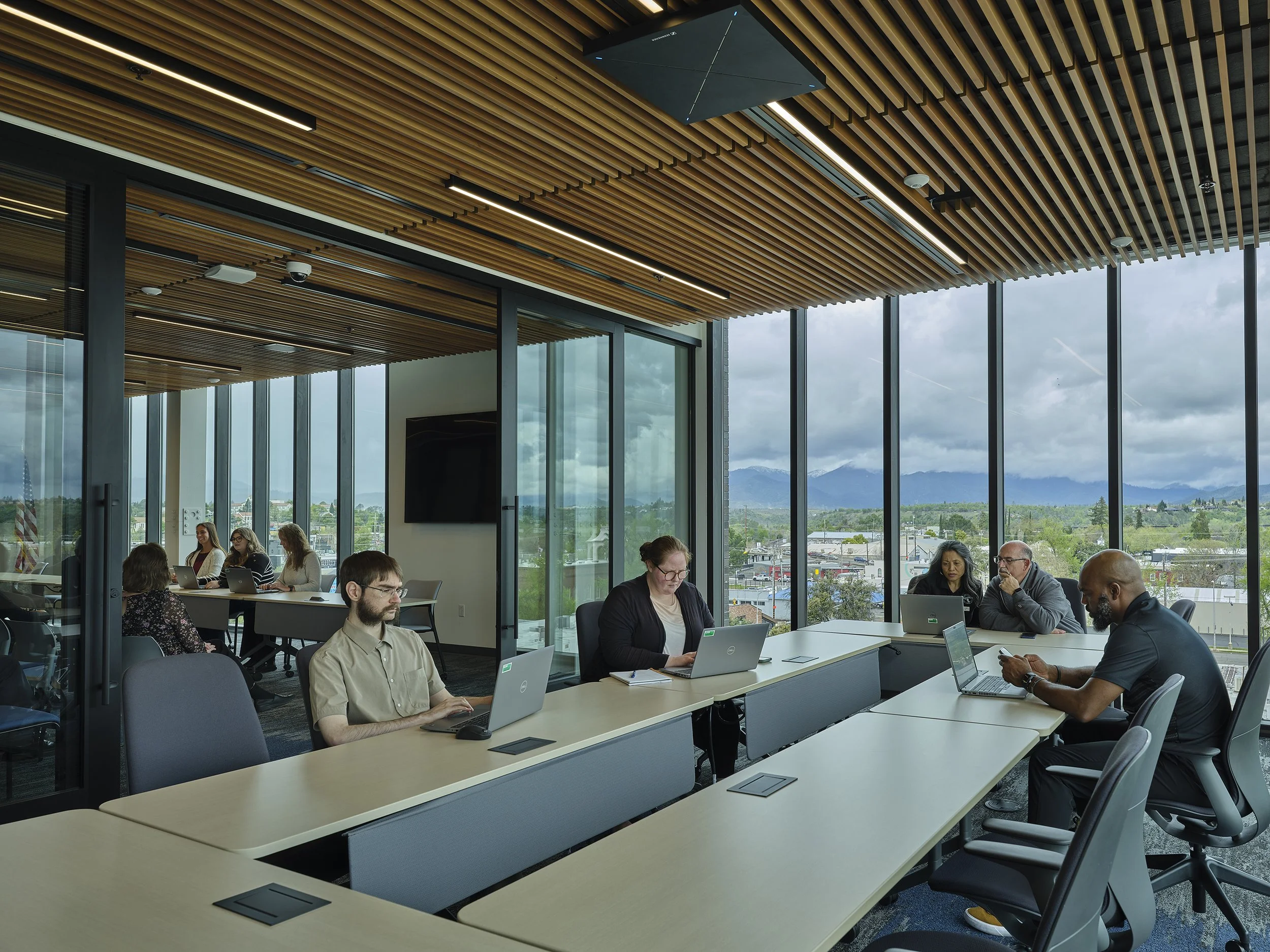 Shasta College in Redding, California. Part of Block 7 Development. People sitting around a conference table working on laptops inside a modern office with large windows showing mountains and a cloudy sky outside.