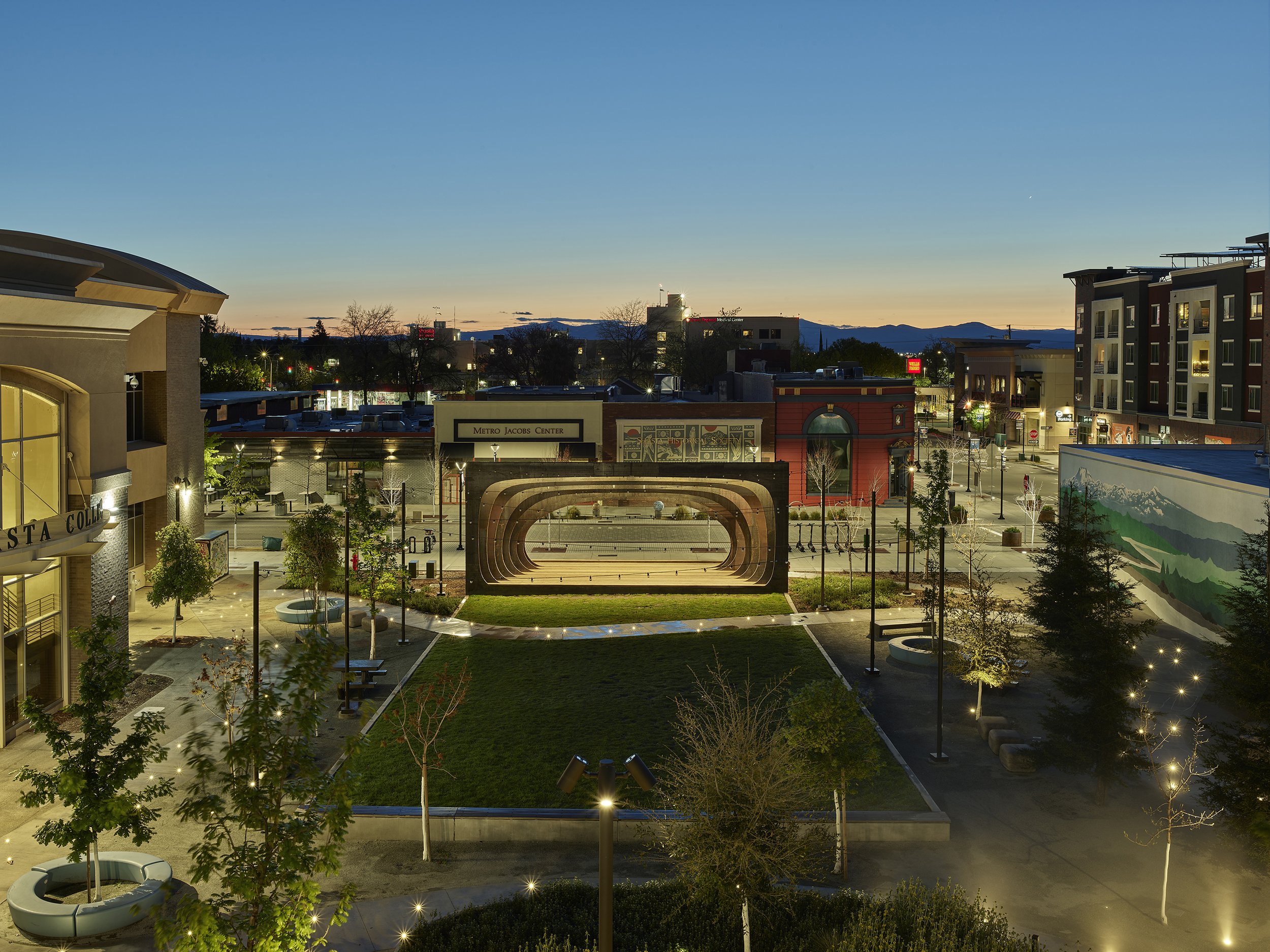 Block 7 Development in Redding, California. Whistle Stop Park custom steel pavilion. A cityscape at dusk with buildings, trees, and a stage or amphitheater in a park, with mountains in the background and a clear sky.
