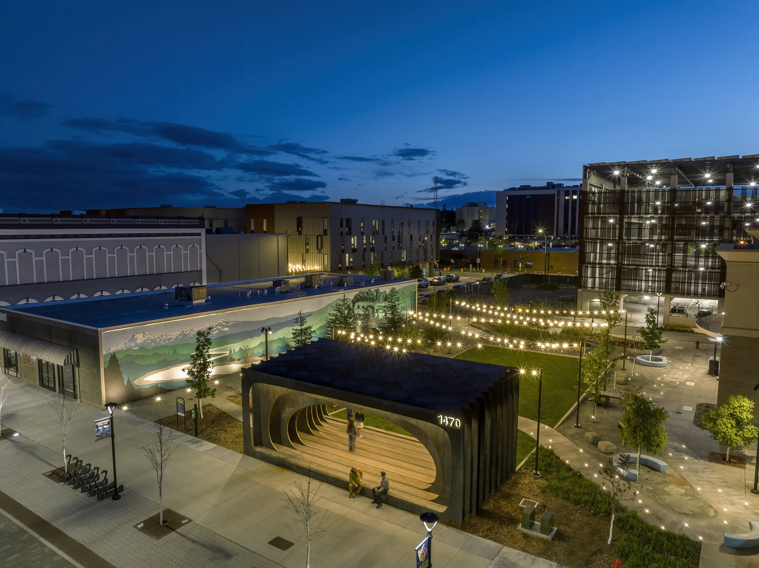 An aerial view of Whistle Stop Park at Block 7 Development in Redding, California. Nighttime view of a modern urban area with illuminated buildings, a mural on a wall, and a small park with trees and benches.