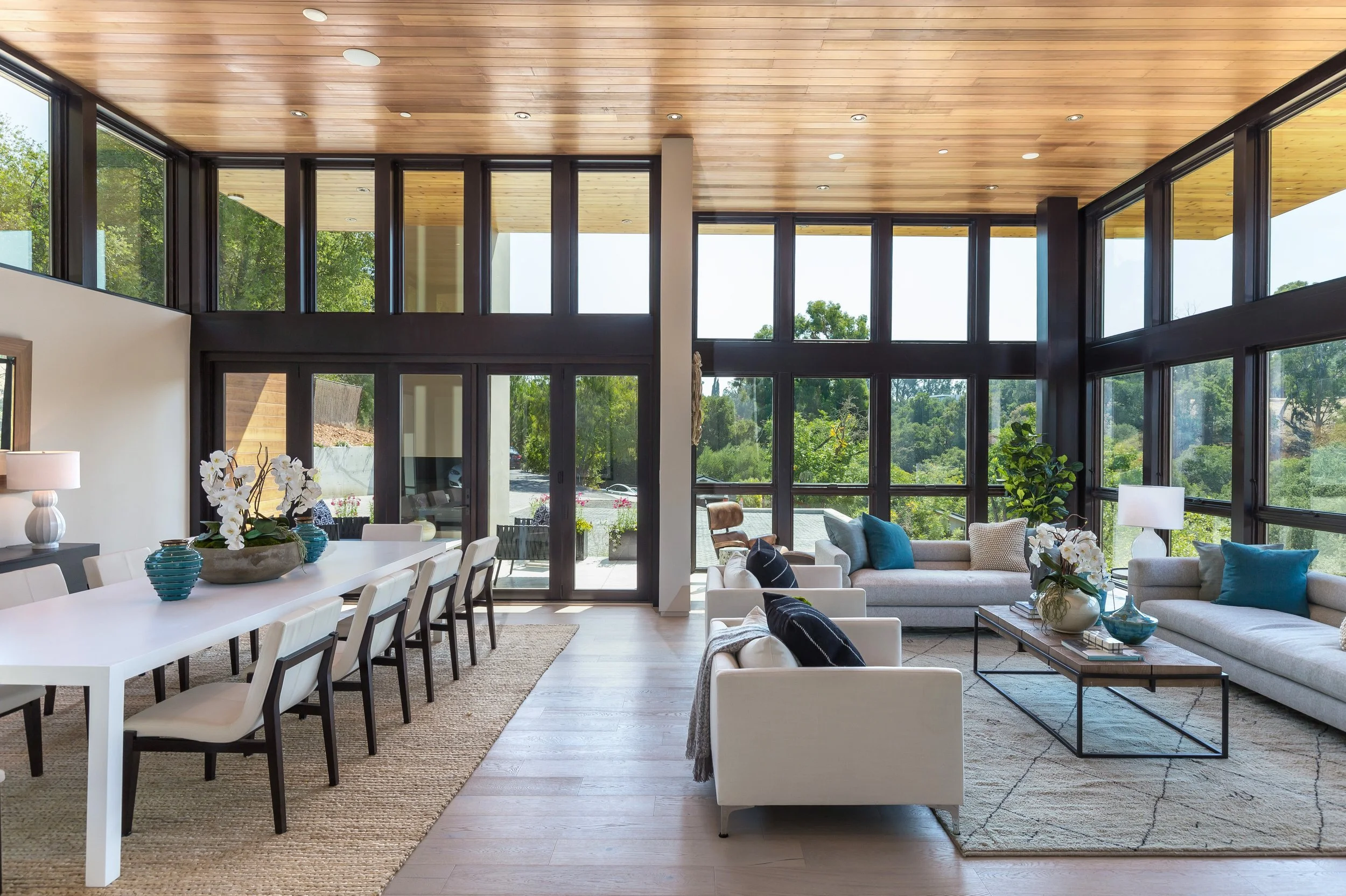 Los Altos Hillside Home in Los Altos, California. Interior of a modern living and dining room with large windows, wooden ceiling, white furniture, potted plants, and view of greenery outside.