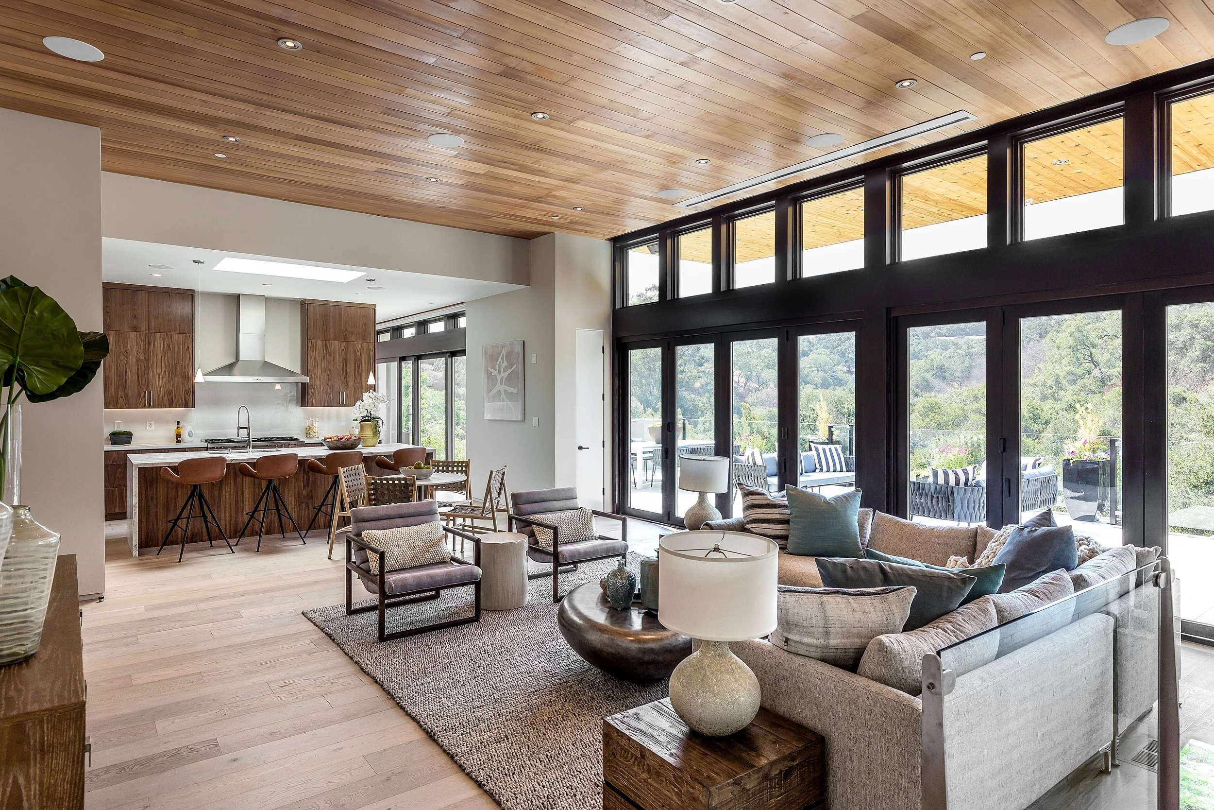 Los Altos Hillside Home in Los Altos, California. Modern open-concept living area with large glass windows, wooden ceiling, kitchen with island, and seating area with sofas and chairs.