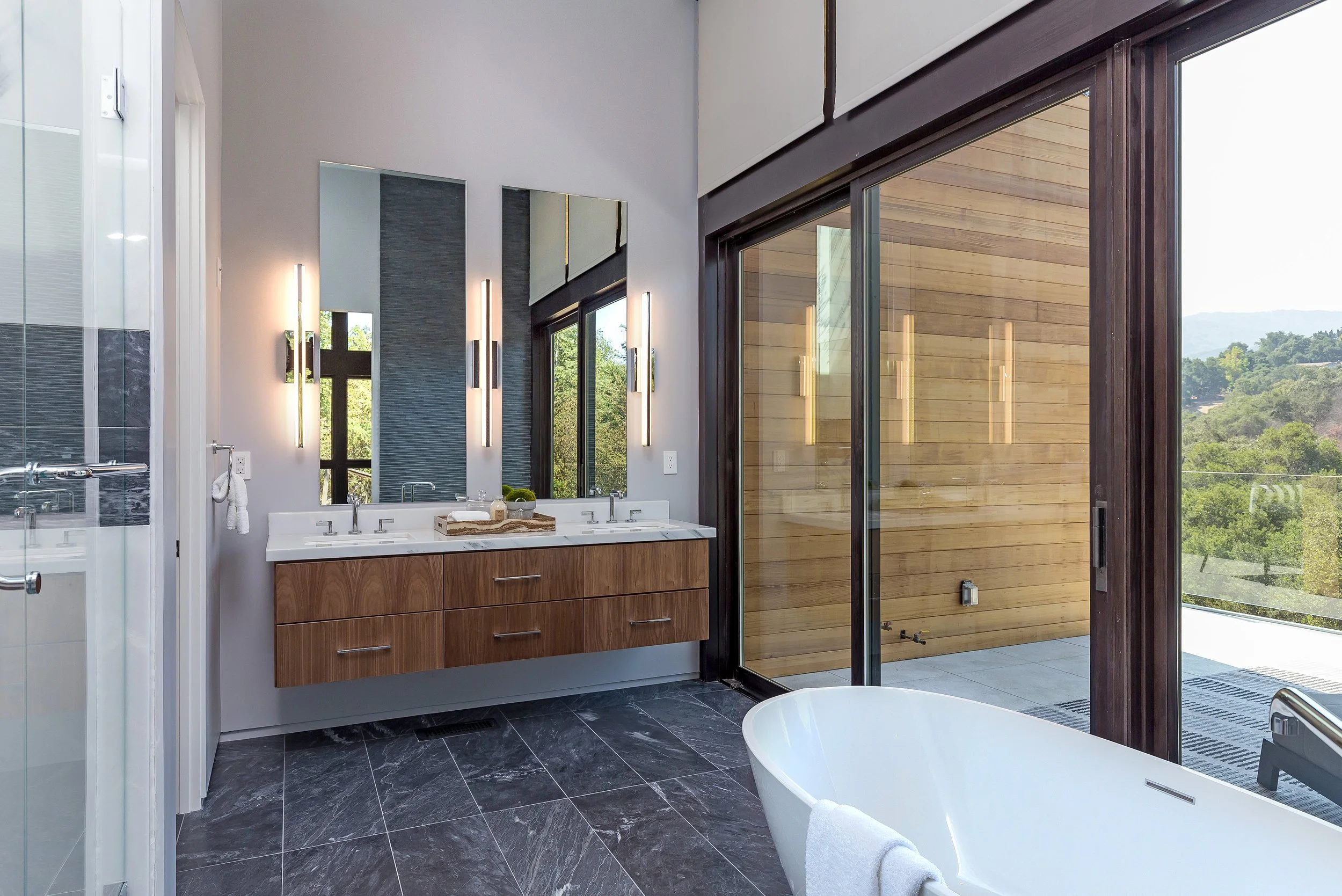 Los Altos Hillside Home in Los Altos, California. Modern bathroom with a white bathtub, wooden vanity with two sinks, and a large glass sliding door leading to an outdoor patio with wooden siding and scenic landscape.