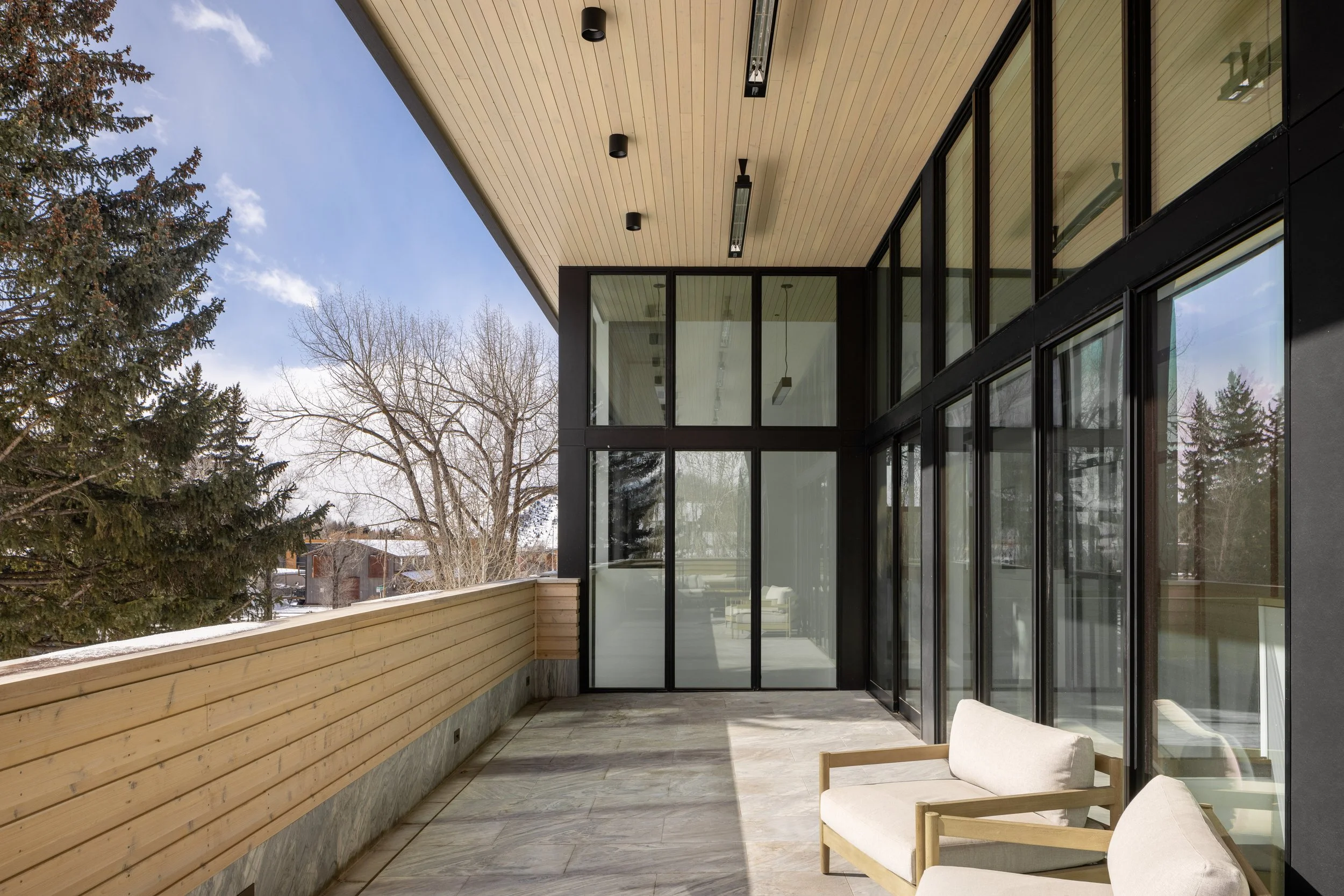 Office at Snow King in Jackson, Wyoming. Modern balcony with wooden railing, large glass windows, and outdoor seating, overlooking snow-covered trees and a partly cloudy sky.