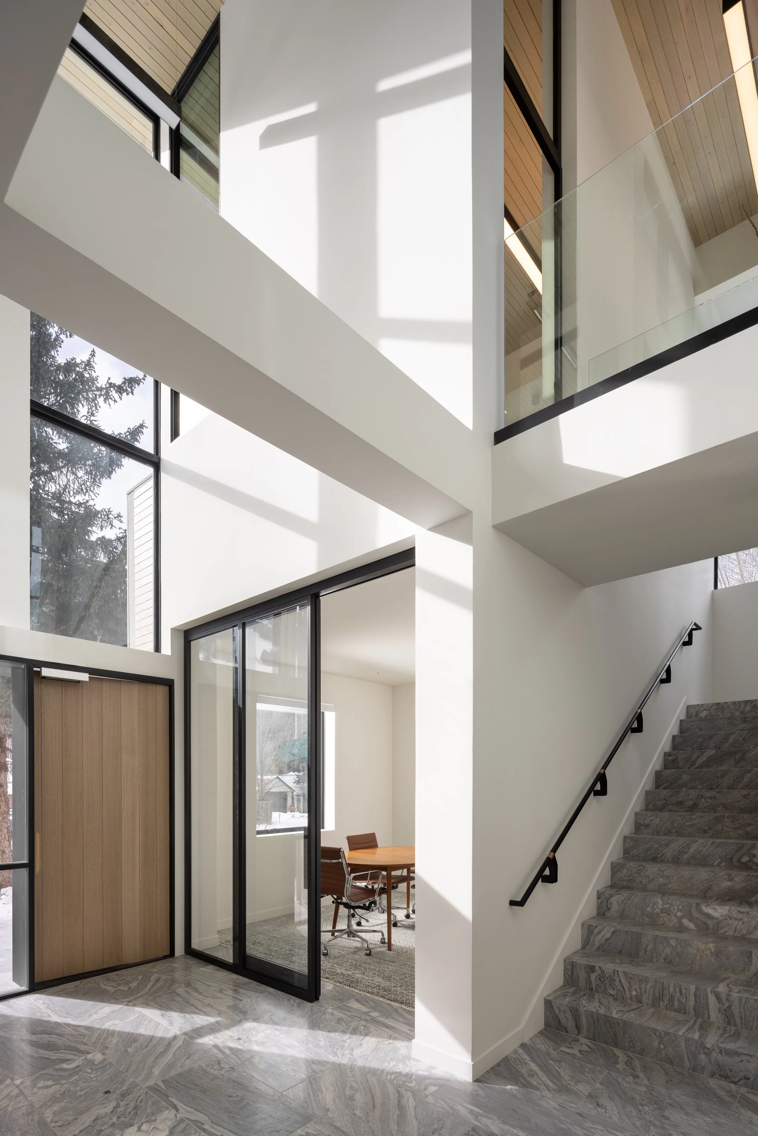 Office at Snow King in Jackson, Wyoming. Interior view of a modern building with marble flooring, large windows, a staircase with grey marble steps and a black handrail, and a glass-enclosed room with a wooden door and chairs inside.