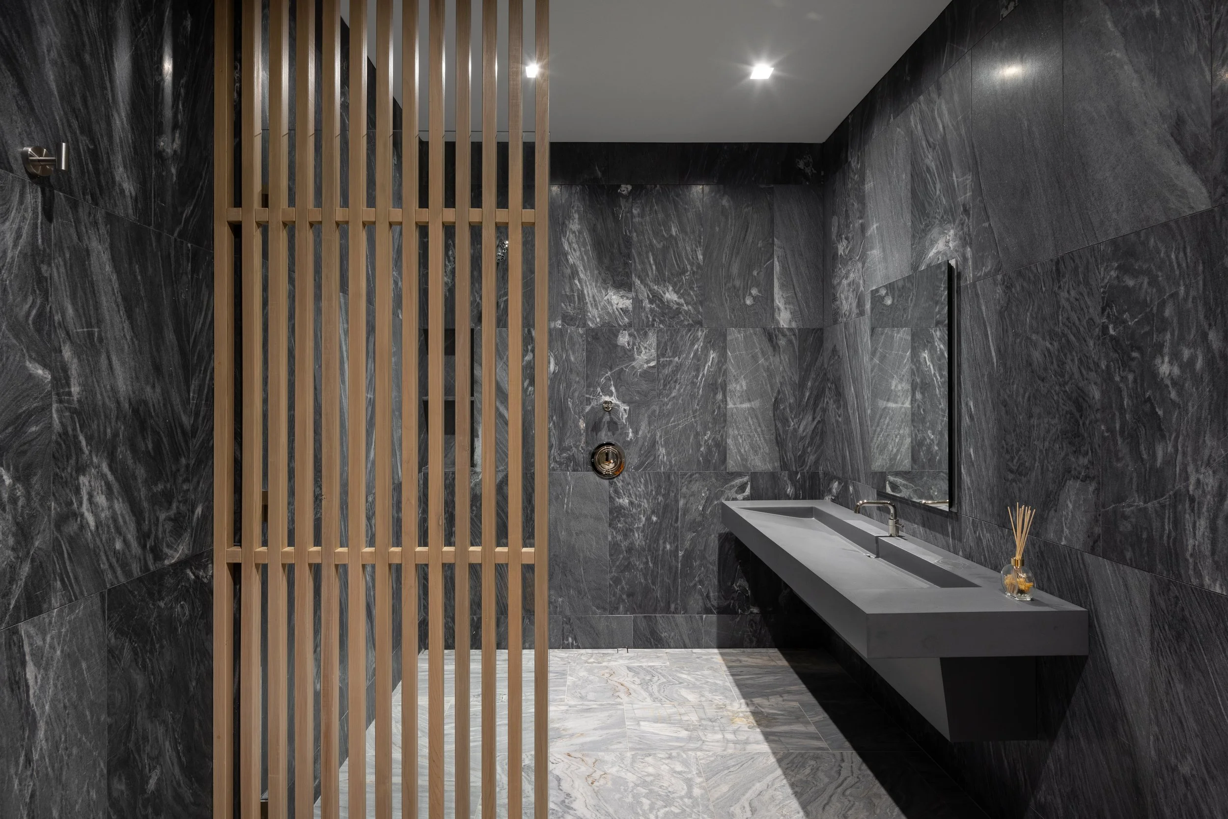 Office at Snow King in Jackson, Wyoming. Modern bathroom with dark gray marble walls and floor, a floating gray sink with a chrome faucet, a mirror, a soap and reed diffuser on the sink, a wooden slat room divider, and ceiling lights.