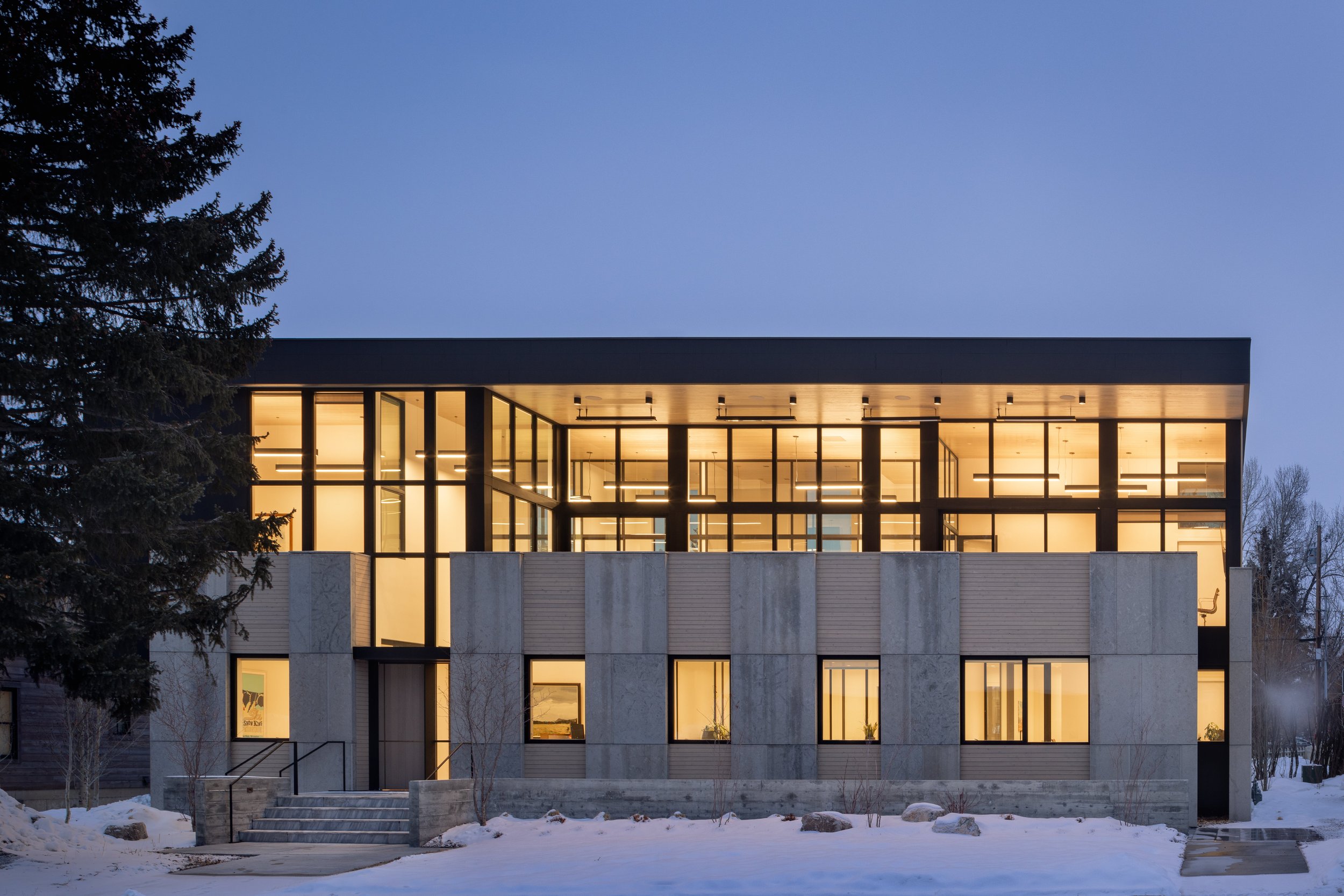 Office at Snow King in Jackson, Wyoming. Modern building with large glass windows and warm interior lighting, surrounded by snow and trees at dusk.