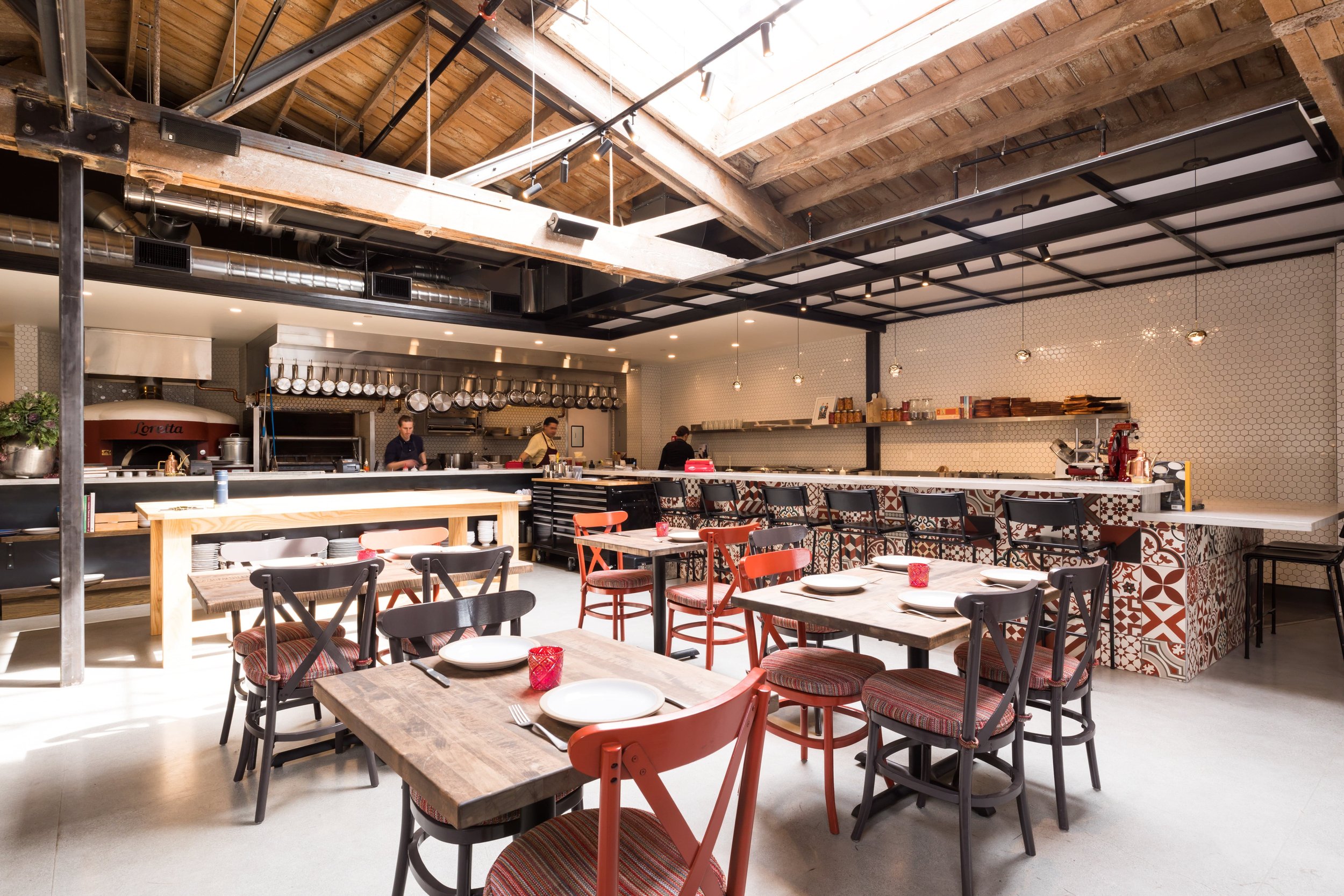 Che Fico restaurant in San Francisco, California. Interior of a modern restaurant with wooden and tiled decor, tables set with plates, glasses, and cutlery, and staff working behind the counter.